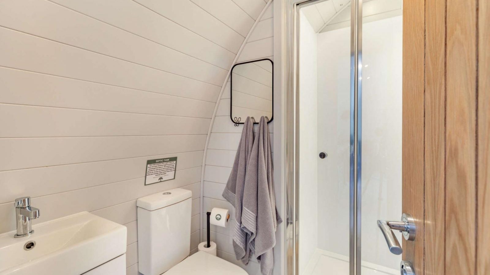 Modern bathroom with toilet, sink, and glass-enclosed shower offers a retreat-like ambiance. A light wood door and white panel walls enhance the serene atmosphere, reminiscent of glamping luxury. Towels hang nearby for convenience.