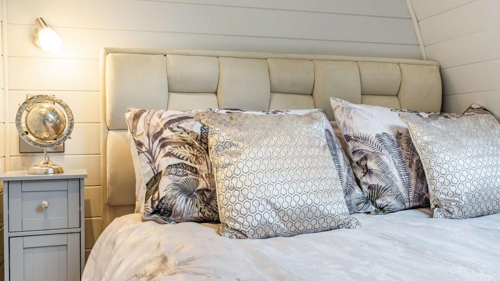 A hidden retreat awaits in the bedroom, featuring a beige tufted headboard, floral-patterned bedding, a nightstand with a globe lamp, and a decorative mirror.