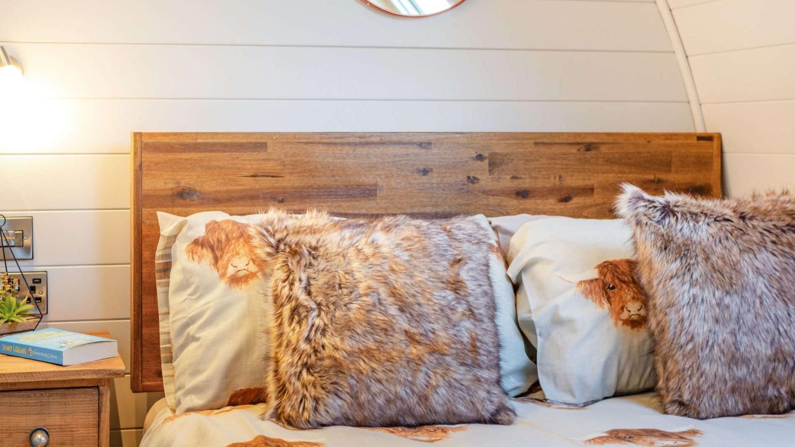 Cozy bed with animal print bedding and fur pillows, complemented by a wooden headboard and a round mirror on a paneled wall. This hidden retreat offers the perfect blend of rustic charm and glamping luxury.