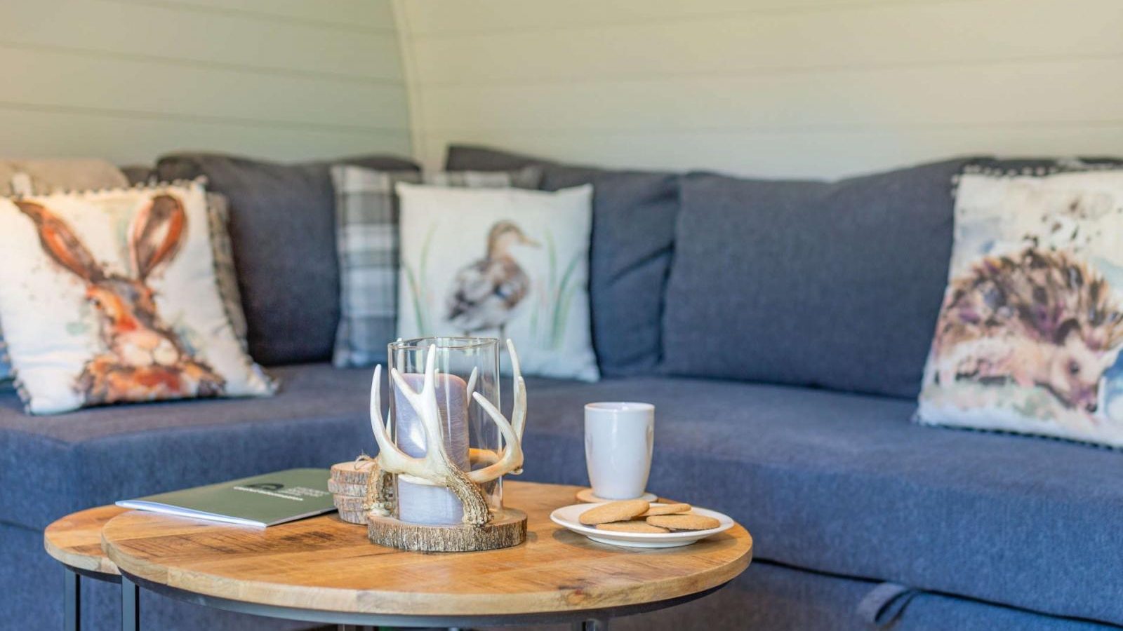 Cozy living room with a gray sofa, nature-themed pillows, and a wooden coffee table reminiscent of a hidden retreat. A candle flickers softly beside a mug and cookies, creating the perfect glamping atmosphere indoors.