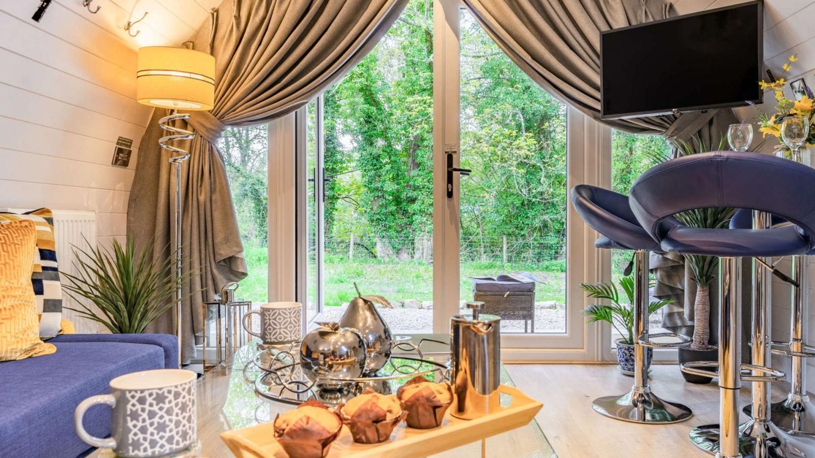 Experience a cozy hidden retreat with a glass door view, stylish furniture, and a wall-mounted TV. Perfect for a touch of glamping, enjoy muffins on a tray alongside a charming teapot in this inviting living area.