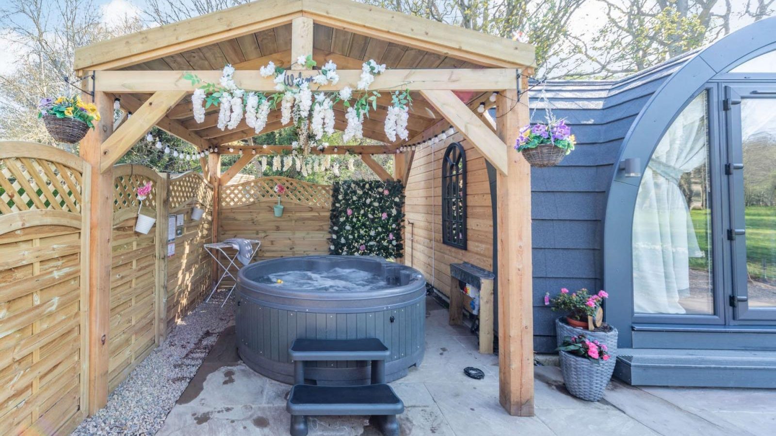 Discover a hidden retreat featuring an outdoor hot tub under a wooden pergola adorned with floral decorations. Nestled next to a charming glamping pod, this serene escape is surrounded by a rustic wooden fence, offering the perfect blend of comfort and nature.