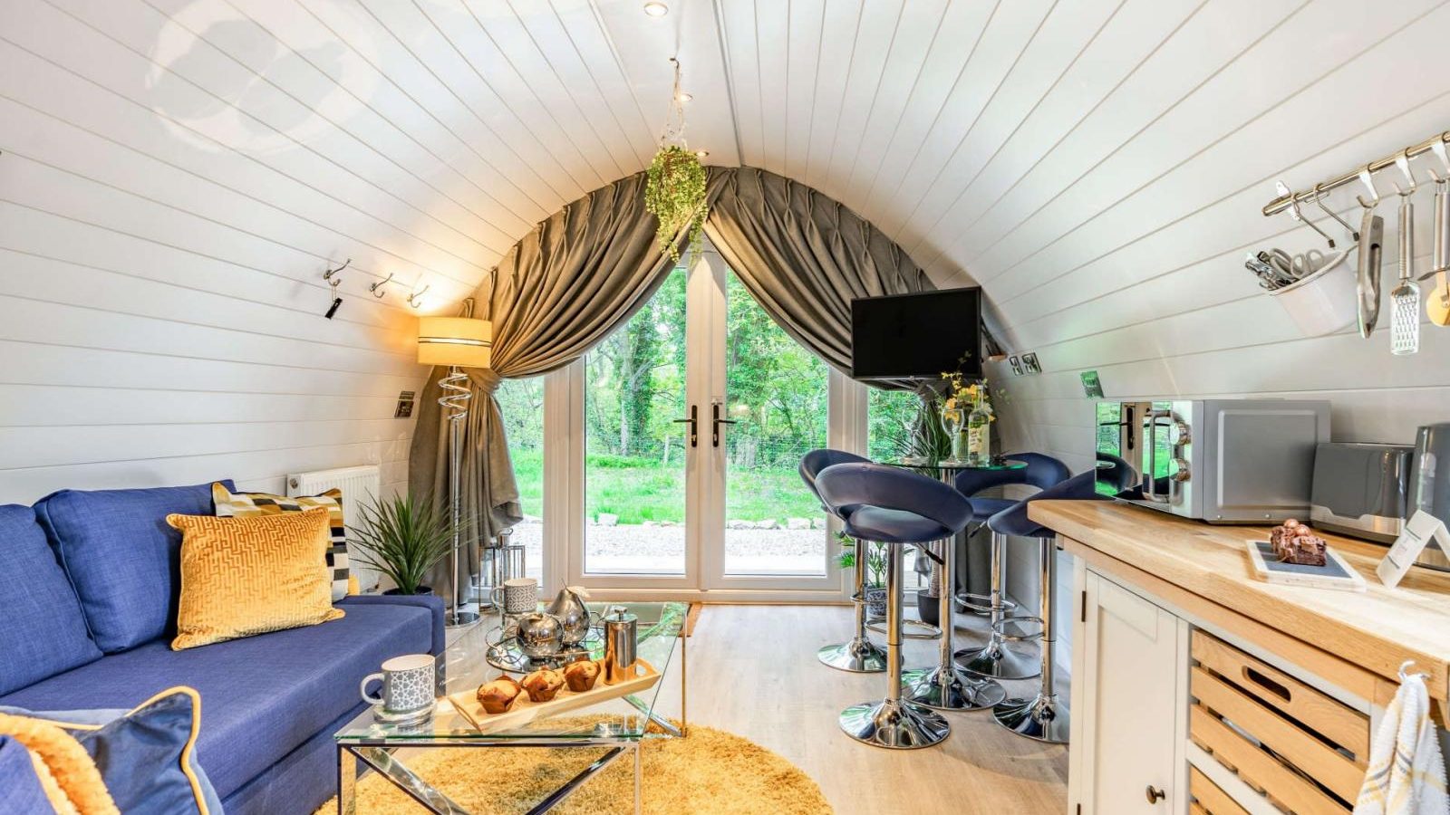 Experience the ultimate retreat in this cozy, modern tiny home featuring a blue sofa, kitchenette with bar stools, large windows, and wooden decor that's perfect for glamping enthusiasts.