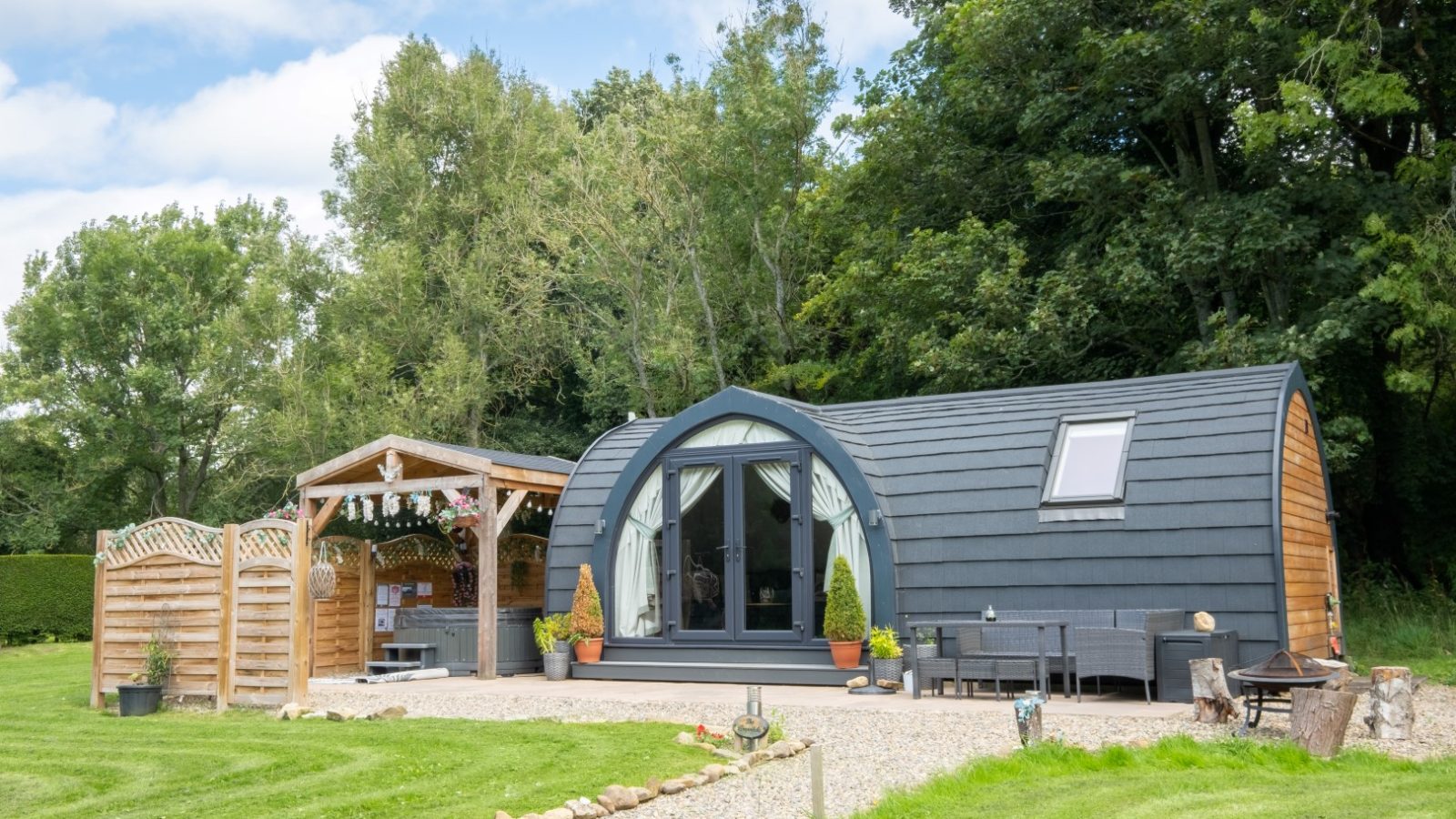 A small, modern cabin with a rounded roof sits as a hidden retreat in a grassy area, surrounded by trees. An outdoor seating area under a pergola invites you to relax and enjoy the serenity of this glamping escape.