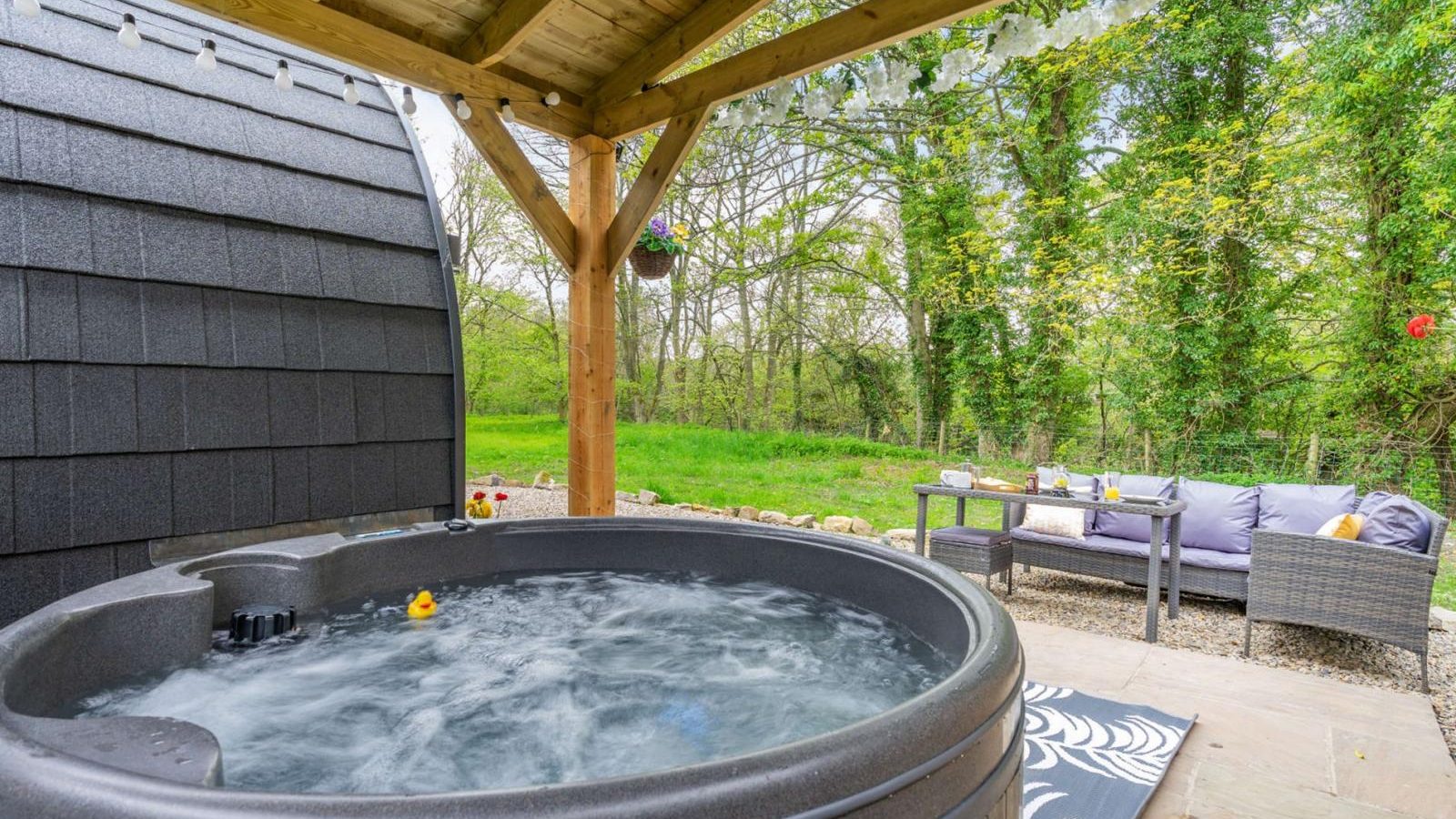 Experience the ultimate glamping escape with an outdoor hot tub under a wooden canopy, offering stunning views of lush greenery. This hidden retreat features a cozy seating area with a gray couch and table, perfect for relaxing in nature's embrace.