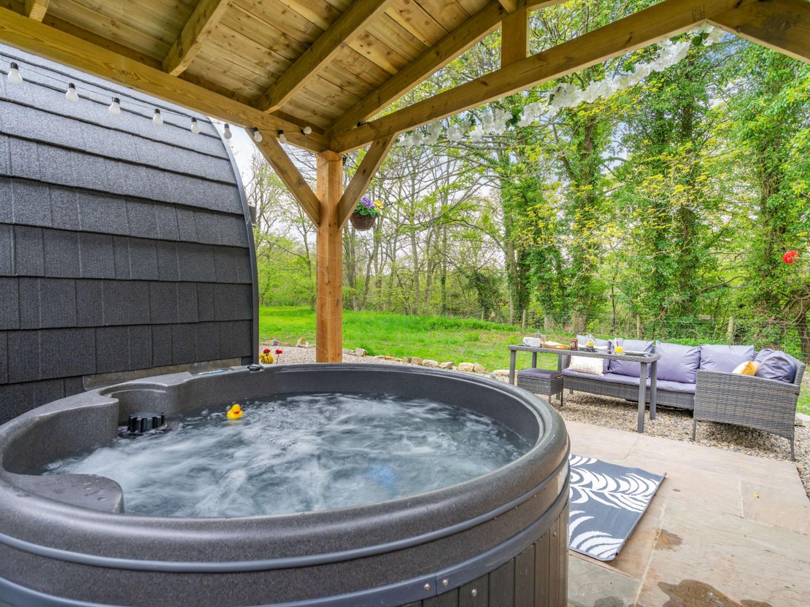 Experience the ultimate glamping escape with an outdoor hot tub under a wooden canopy, offering stunning views of lush greenery. This hidden retreat features a cozy seating area with a gray couch and table, perfect for relaxing in nature's embrace.