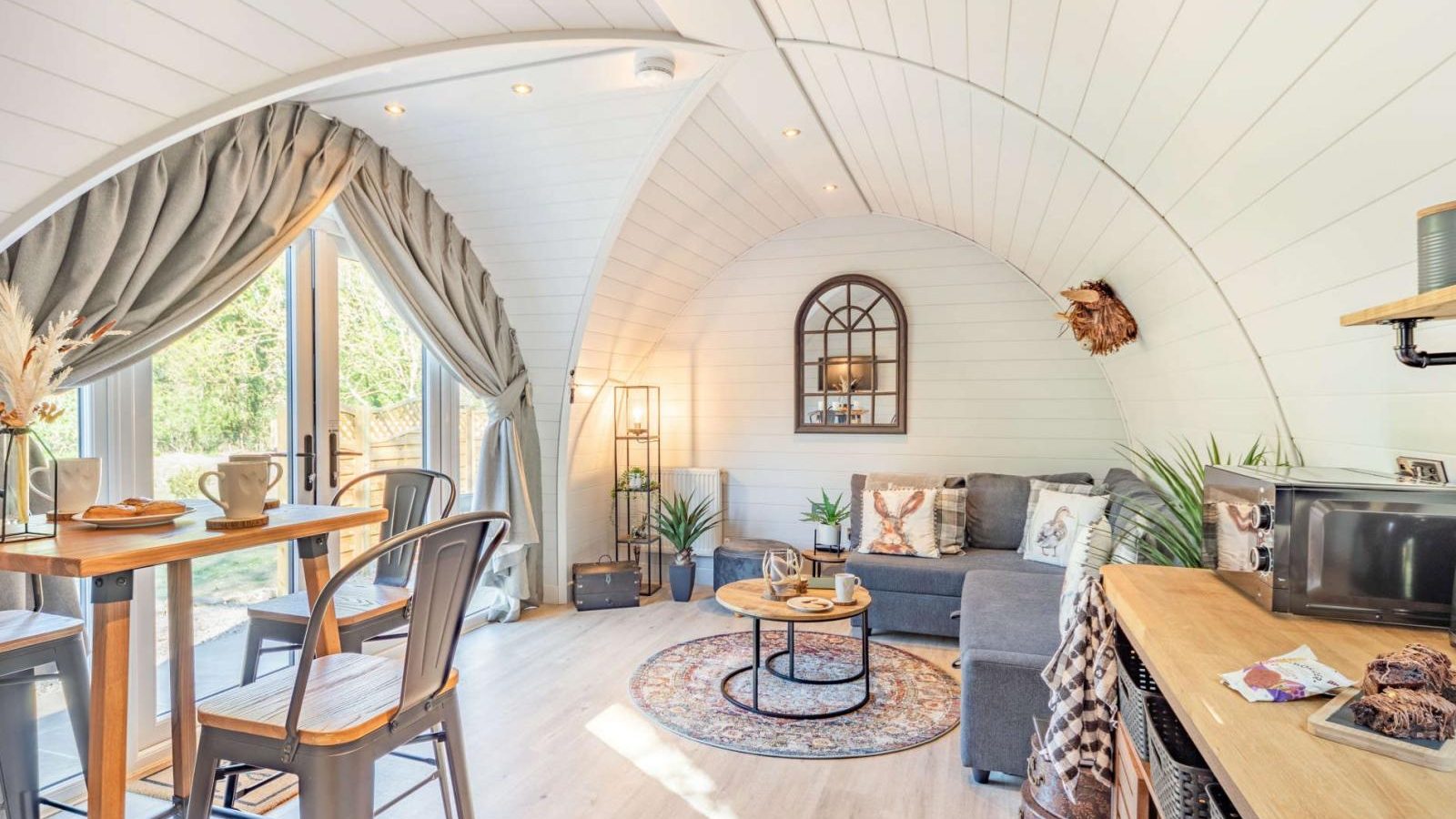 Cozy living space with an arched ceiling, gray sofa, round rug, dining table, and plants. This hidden retreat features a large window draped with curtains, perfect for bringing the outdoors in.