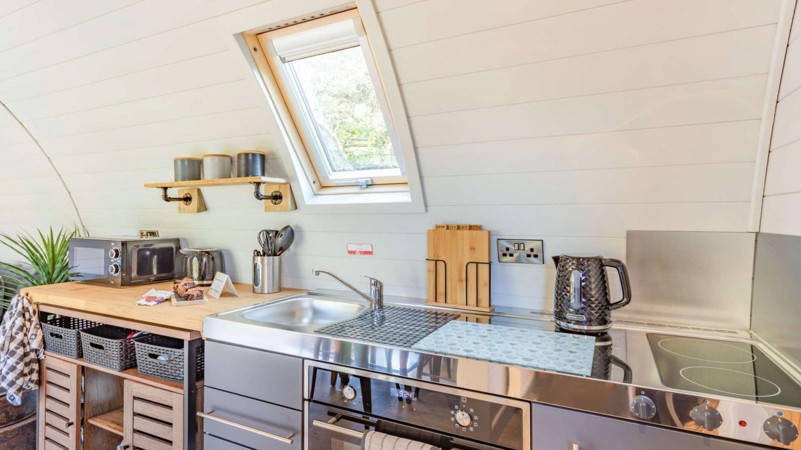 This compact kitchen, perfect for a hidden retreat, features a metal countertop with a sink, stove, and shelves under a skylight window. Cups and toaster sit on the counter, inviting you to enjoy your glamping culinary adventure.