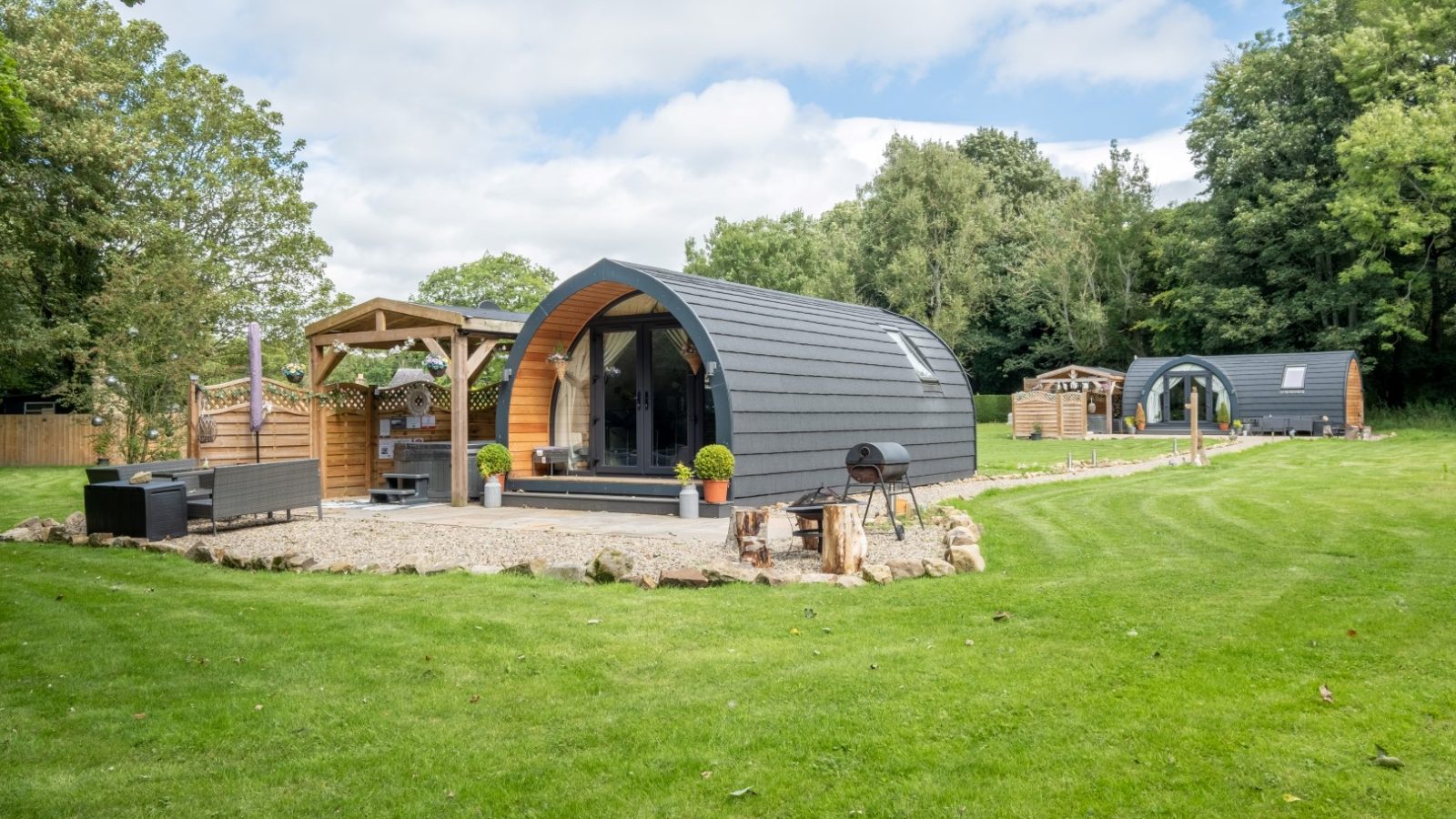 Two modern pod cabins with wooden accents, nestled in a hidden retreat surrounded by lush greenery and clear skies, set on a well-maintained lawn perfect for glamping enthusiasts.