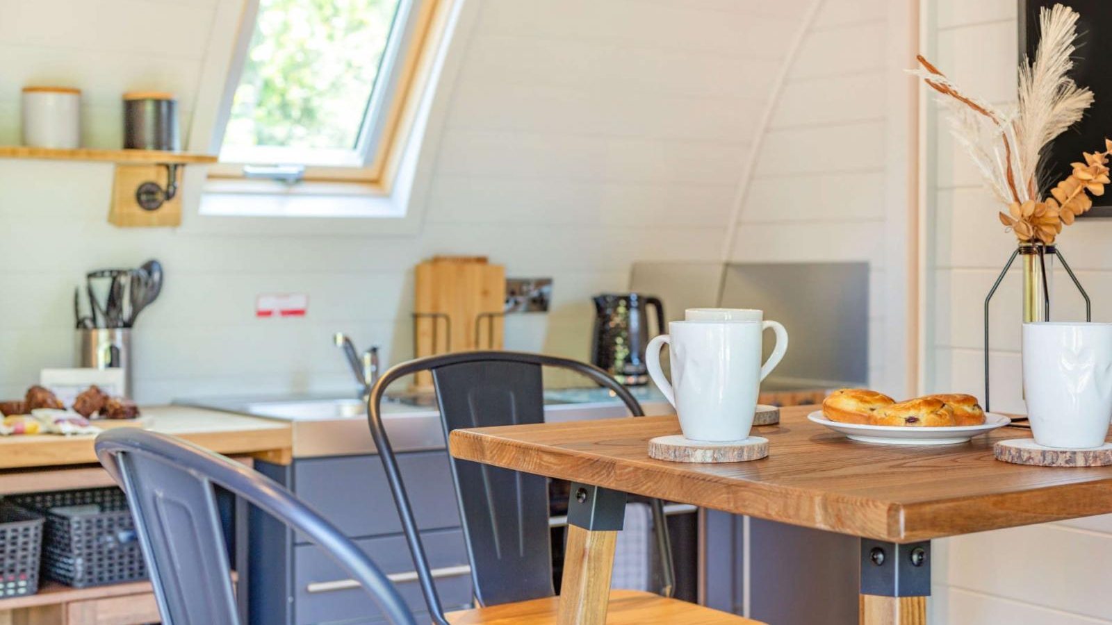 A hidden retreat awaits in this small modern kitchen, featuring a wooden table set with mugs and pastries, metal chairs, and a window welcoming natural light.