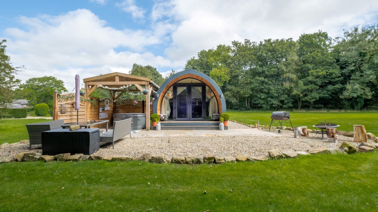 Discover a hidden retreat in this small arched cabin, featuring patio furniture, a gazebo, and a barbecue grill nestled in a grassy yard surrounded by trees—perfect for a glamping getaway.