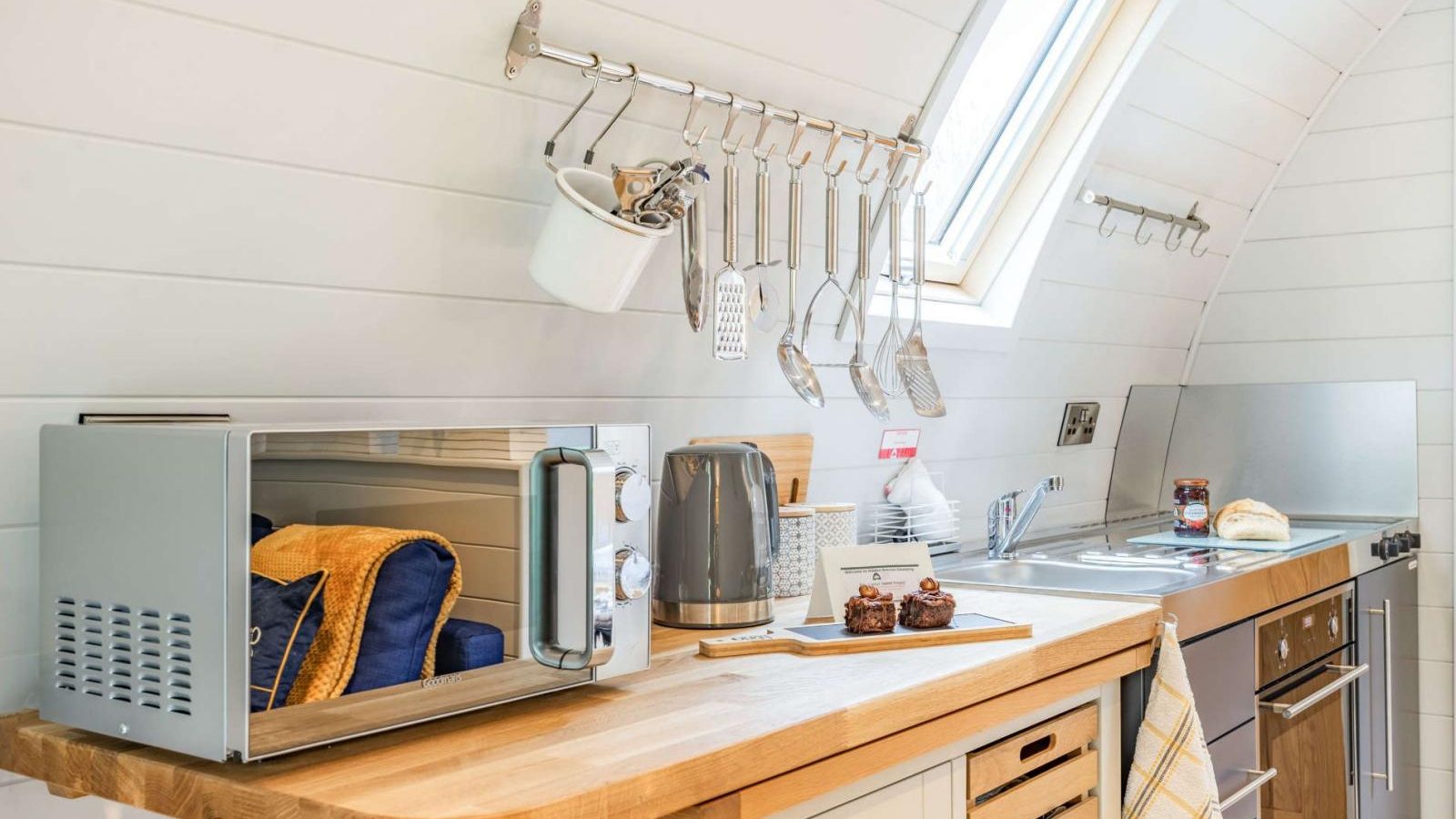 Compact kitchen with wooden countertop, perfect for a hidden retreat, featuring a microwave, kettle, utensils on a rack, and a skylight.