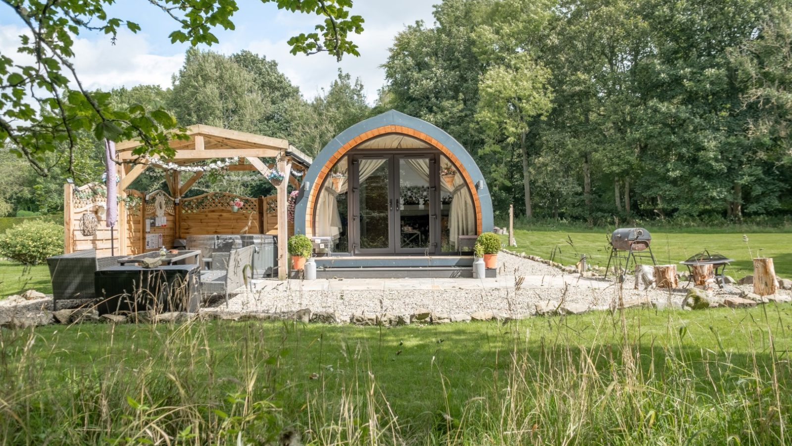 A hidden retreat, this small wooden cabin with a curved roof nestles in a lush green landscape, featuring a deck and outdoor seating area perfect for glamping.