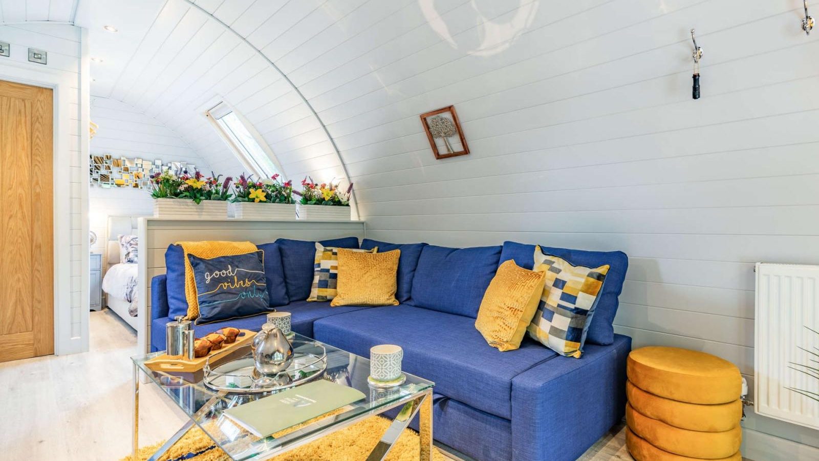 Cozy living room with a blue sofa, yellow pillows, glass coffee table, and floral decorations, creating a hidden retreat. Bedroom visible in the background.