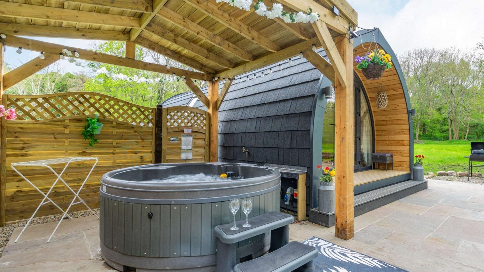 Discover a hidden retreat in this outdoor spa area featuring a wooden shelter, hot tub, steps, and a cozy rug. The pod-like building nestled amid lush greenery offers the perfect glamping experience.