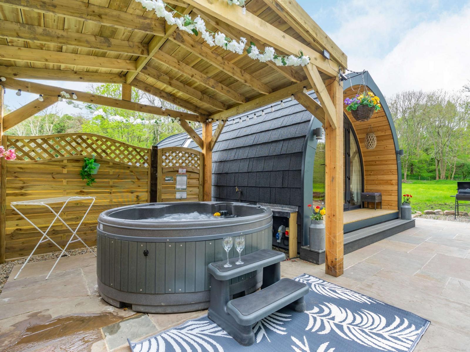 Discover a hidden retreat in this outdoor spa area featuring a wooden shelter, hot tub, steps, and a cozy rug. The pod-like building nestled amid lush greenery offers the perfect glamping experience.