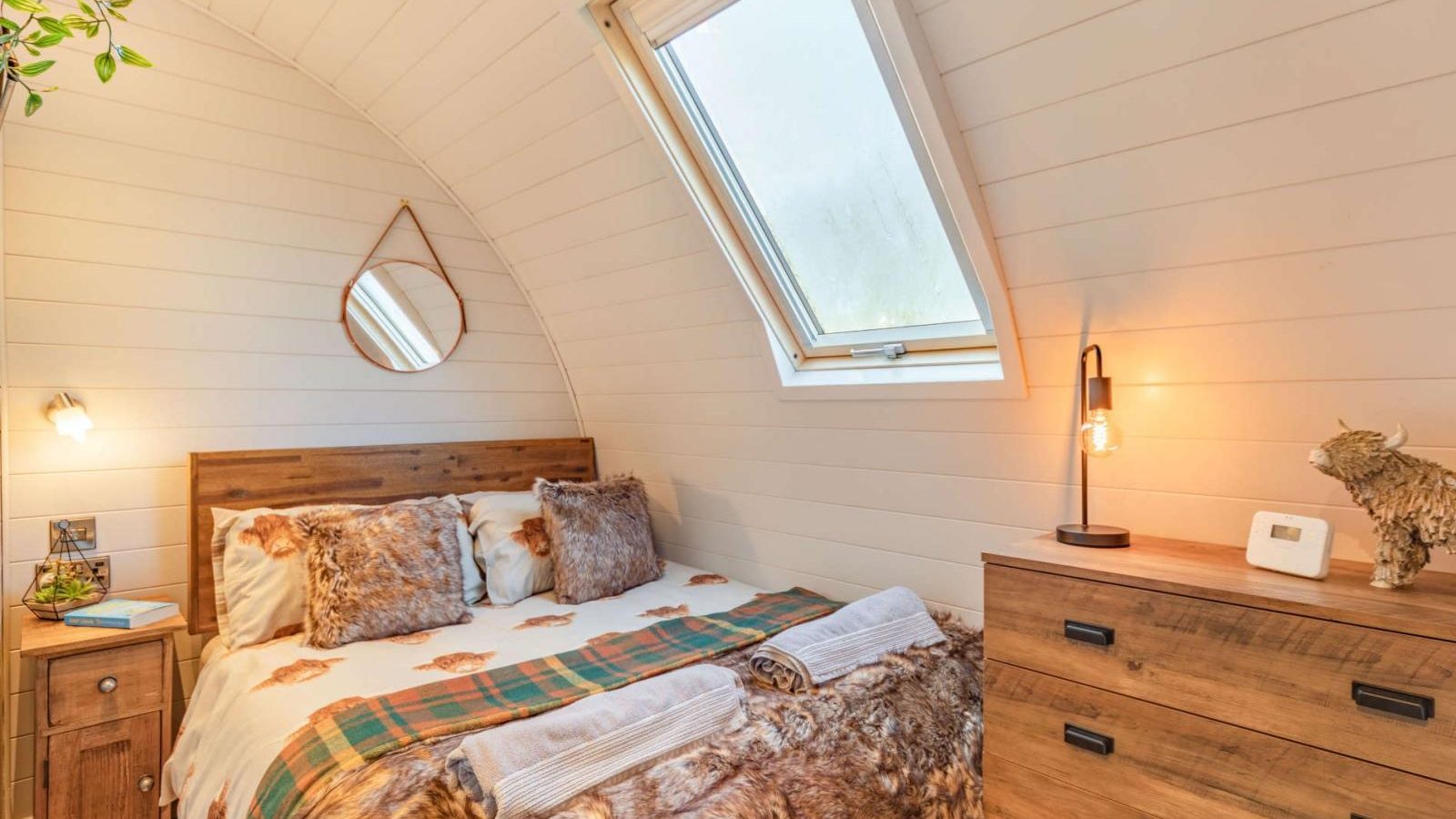 Cozy hidden retreat featuring wood paneling, a skylight, and a double bed with furry blankets. Perfect for glamping, it includes a bedside table, dresser, and wall mirror.