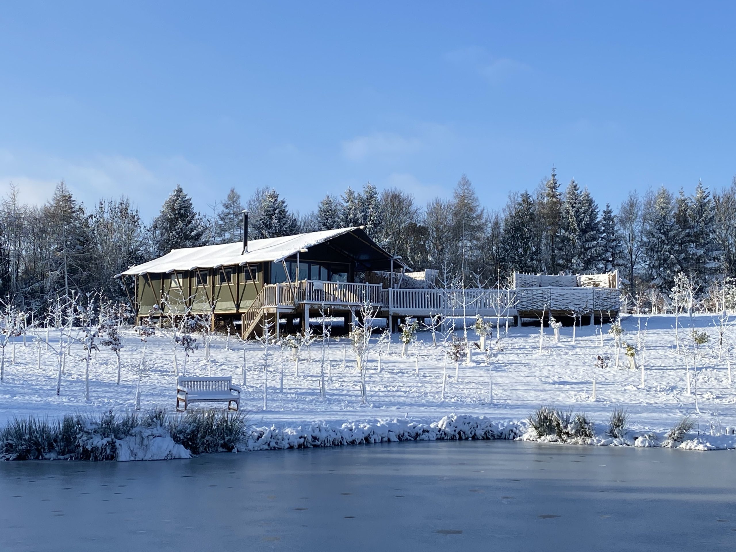 Experience a magical Christmas family break in the UK at this snow-covered cabin. Nestled among trees with a wooden deck, it overlooks a partially frozen pond under a clear blue sky—perfect for making cherished holiday memories.