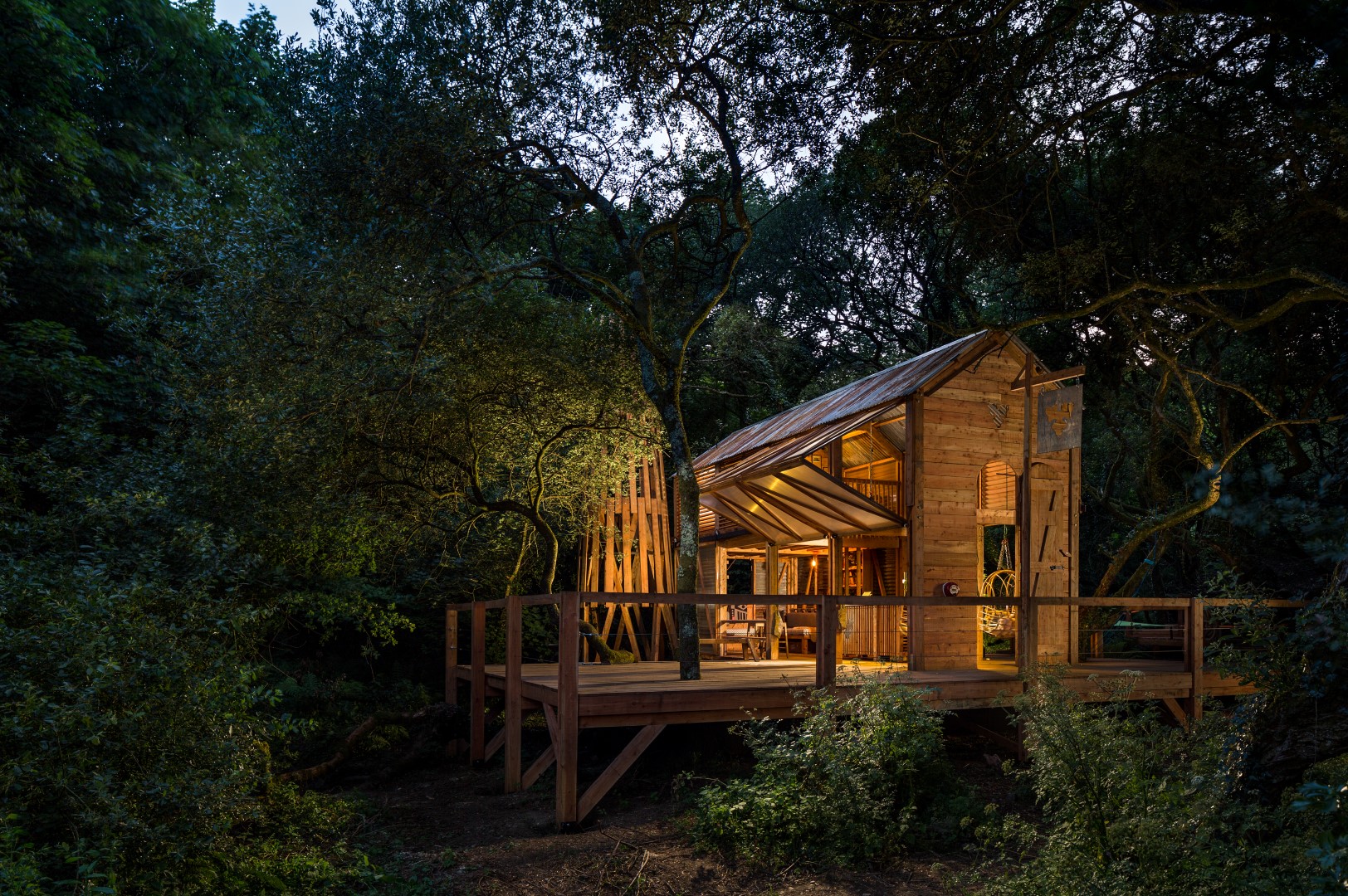 A wooden cabin with large windows and a deck, surrounded by dense trees, softly illuminated in a forest setting at dusk, offers an idyllic retreat reminiscent of Coastal Cornwall breaks in the UK.