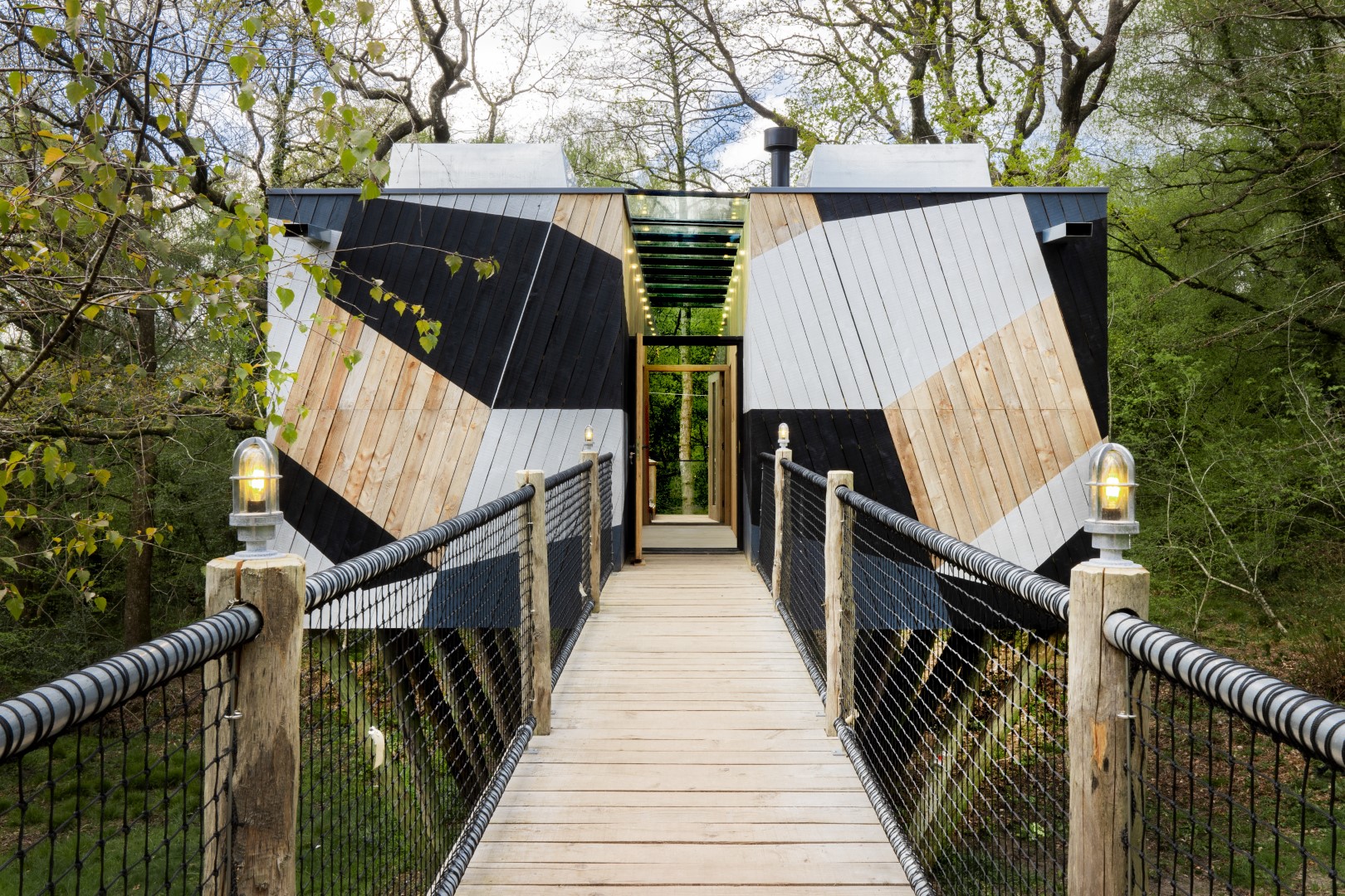 A luxurious wooden bridge ushers you to a modern, geometric house with black, white, and wood panels nestled among UK trees. Perfect for short breaks.