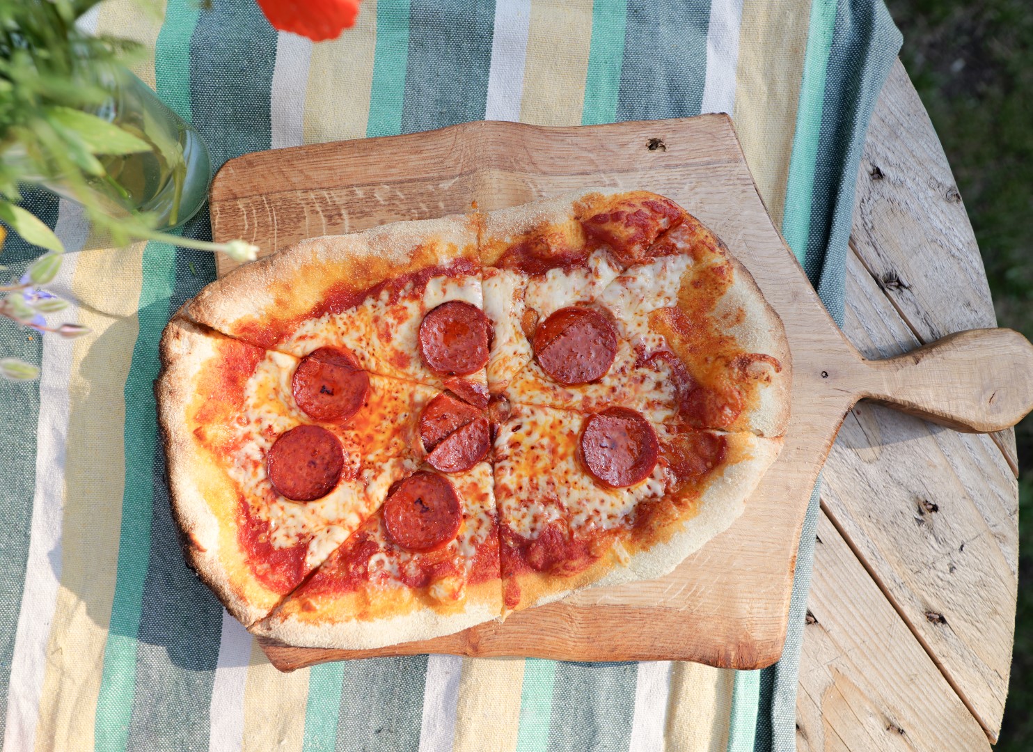 A foodie’s delight: a pepperoni pizza on a wooden board sits enticingly on a striped tablecloth, while nearby, a vase with flowers adds a touch of elegance.