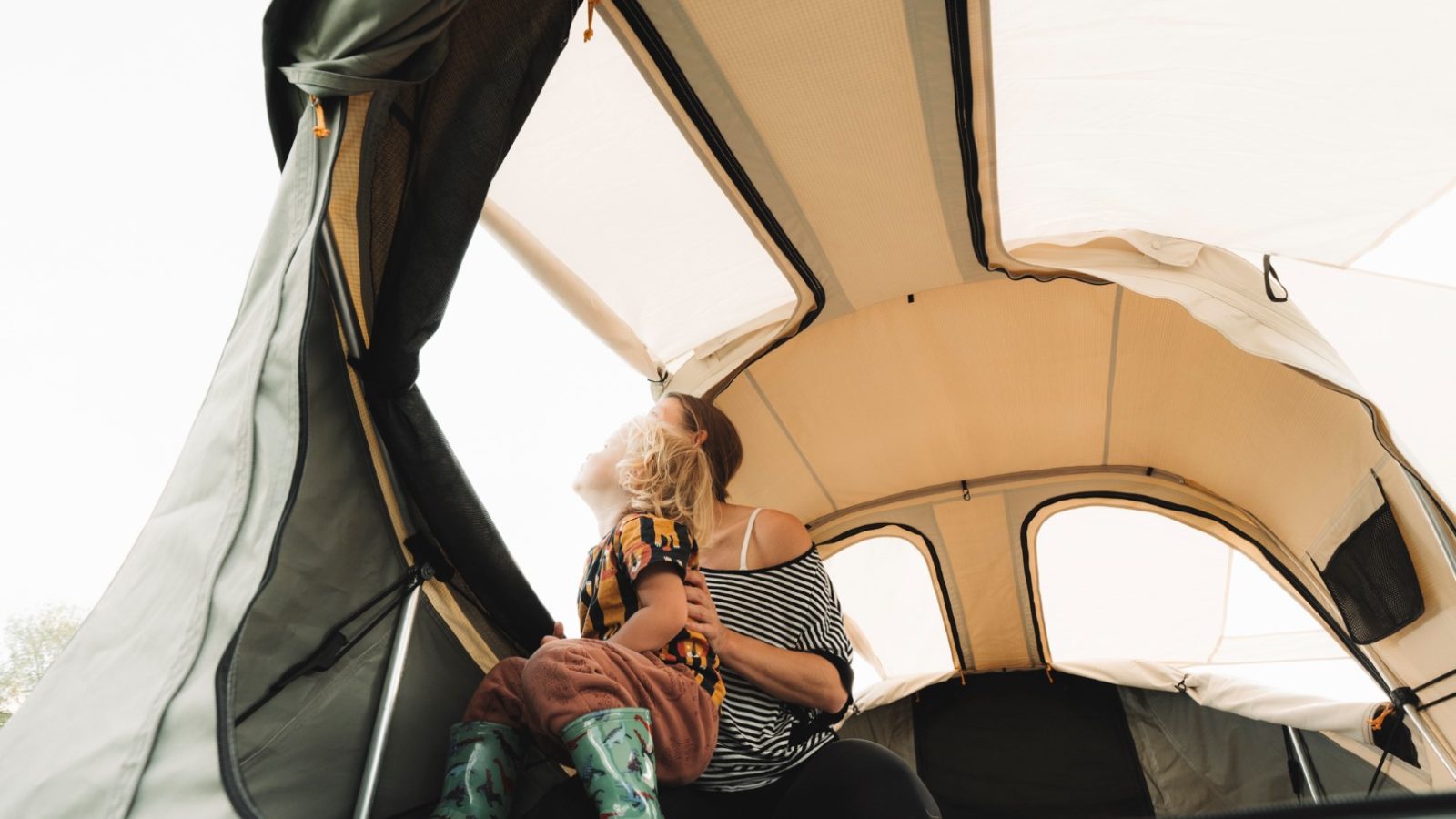 A child and adult sit cozily in a partially open LandyCampers tent, the child gazing upwards while the adult gently holds them from behind.