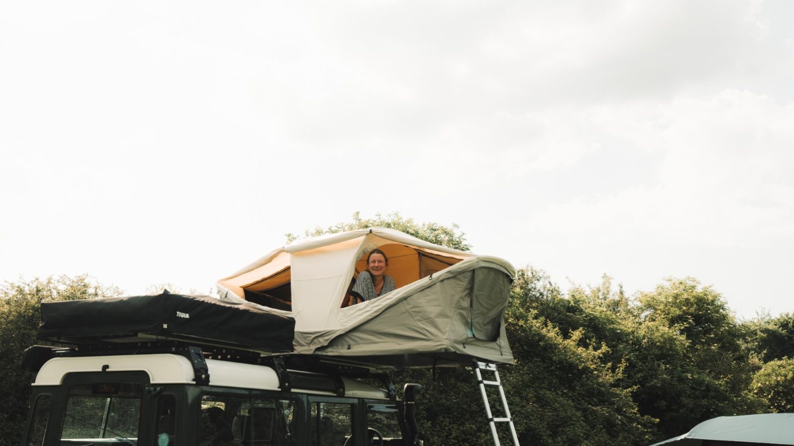 A person smiles from a LandyCampers roof tent perched atop their vehicle, with a ladder leading down, surrounded by lush greenery under a cloudy sky.