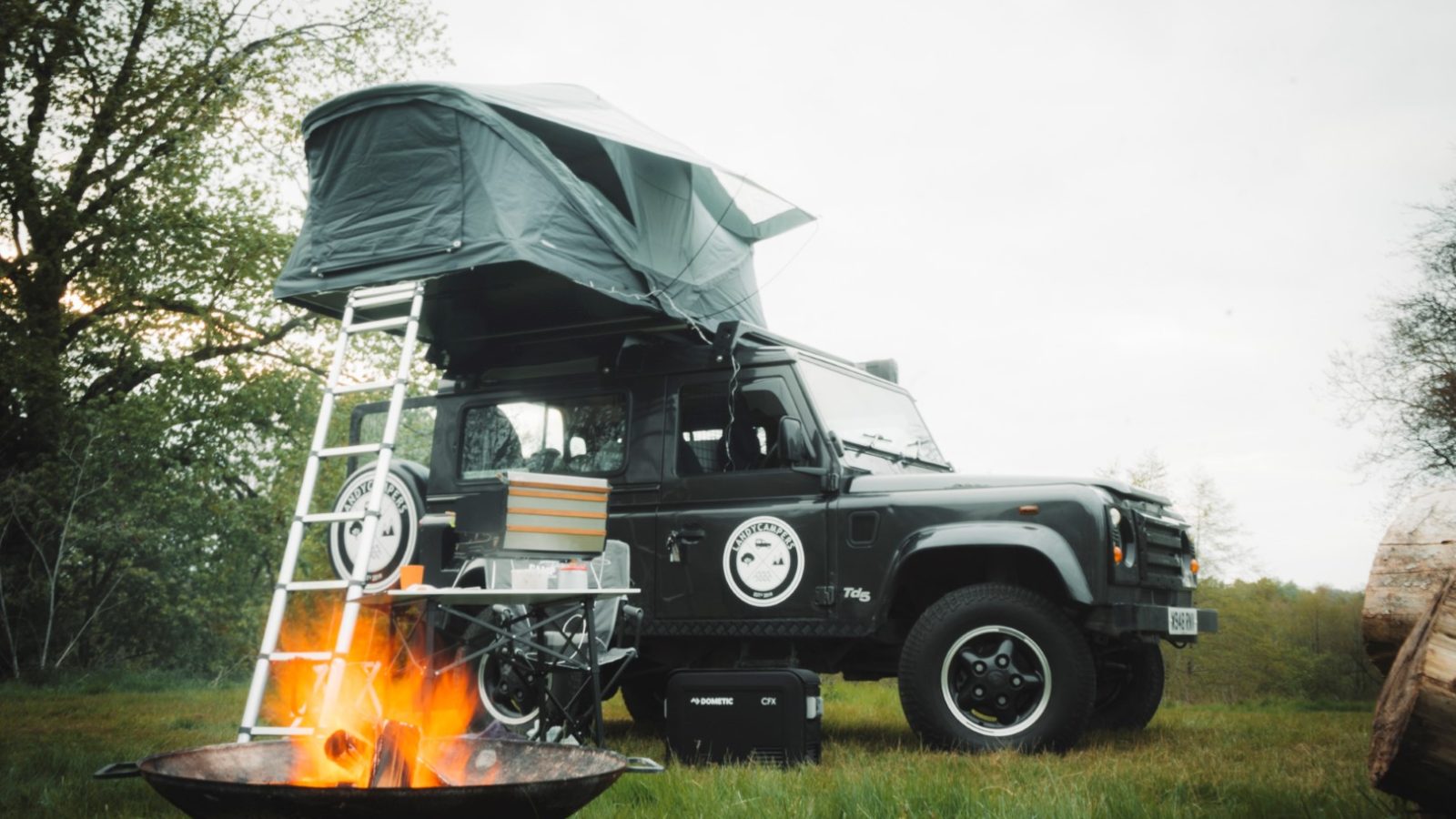 A black off-road vehicle from LandyCampers, equipped with a roof tent, is parked near a crackling campfire in a grassy area surrounded by towering trees.