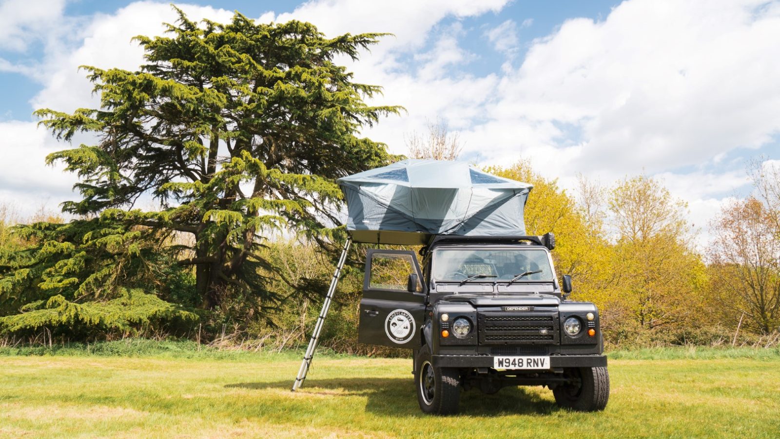 A LandyCampers off-road vehicle with a rooftop tent rests on the grass near a large tree under a partly cloudy sky.