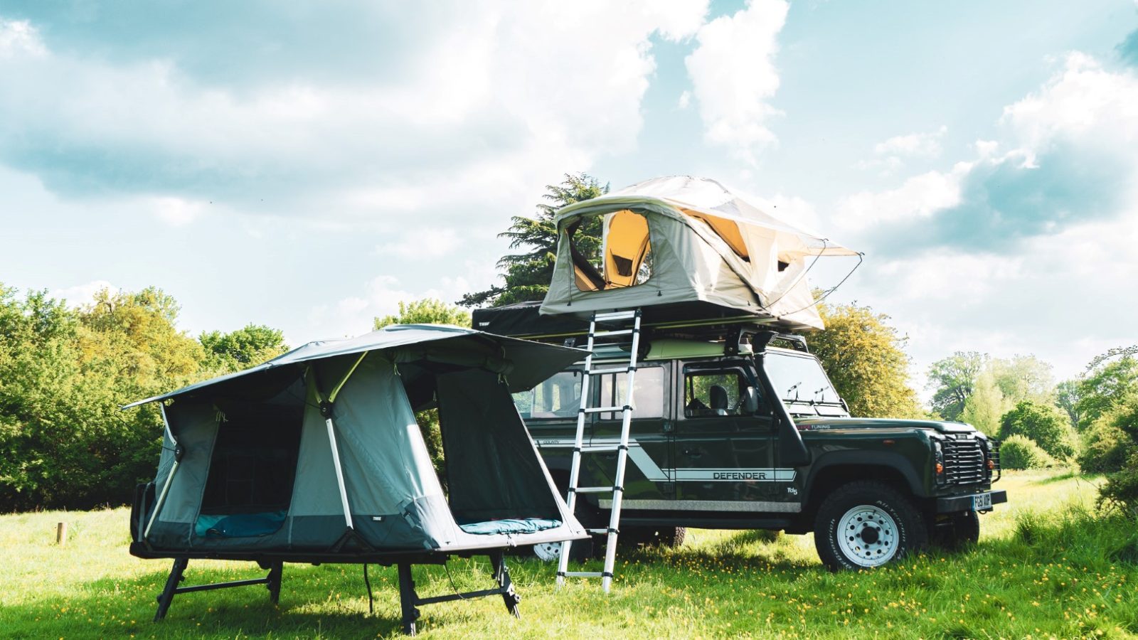 A Land Rover from LandyCampers, equipped with a rooftop tent and accompanied by a foldable tent nearby, sits serenely on a grassy field under a partly cloudy sky.