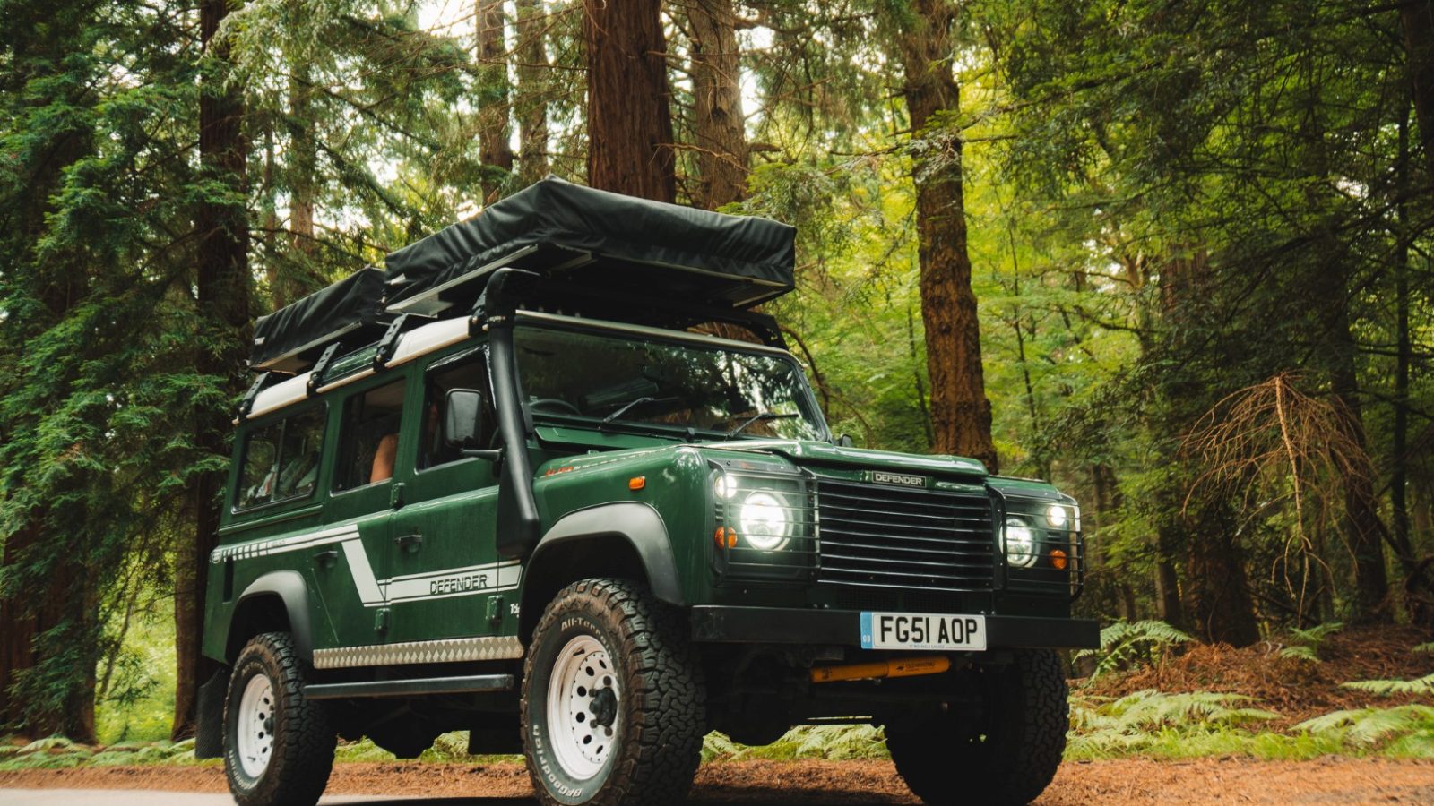 A LandyCampers green off-road vehicle with a roof tent is parked on a forest road, surrounded by tall trees.