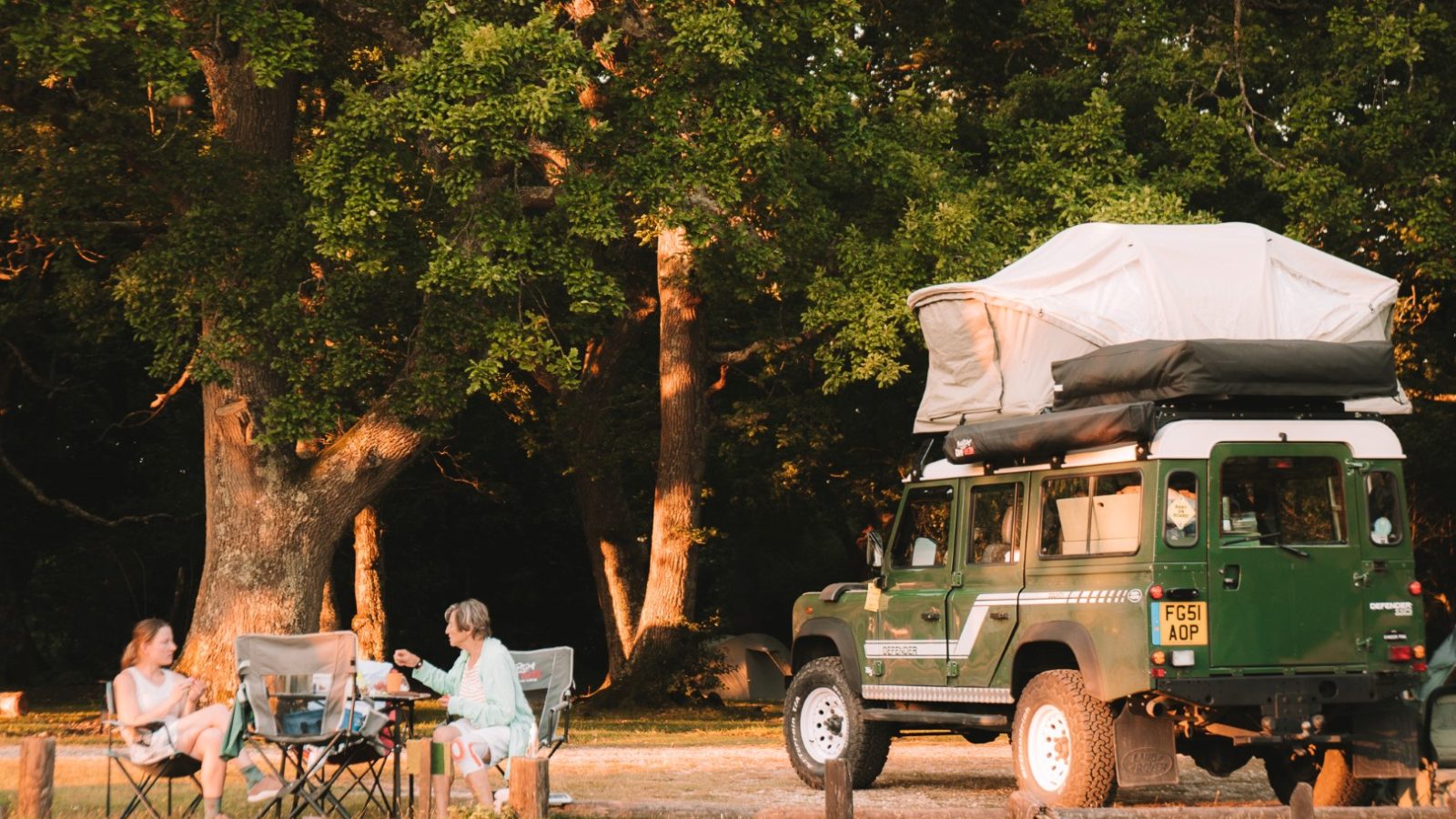 Two people sit on chairs near a green LandyCampers vehicle with a rooftop tent, parked by trees in a sunlit campsite.