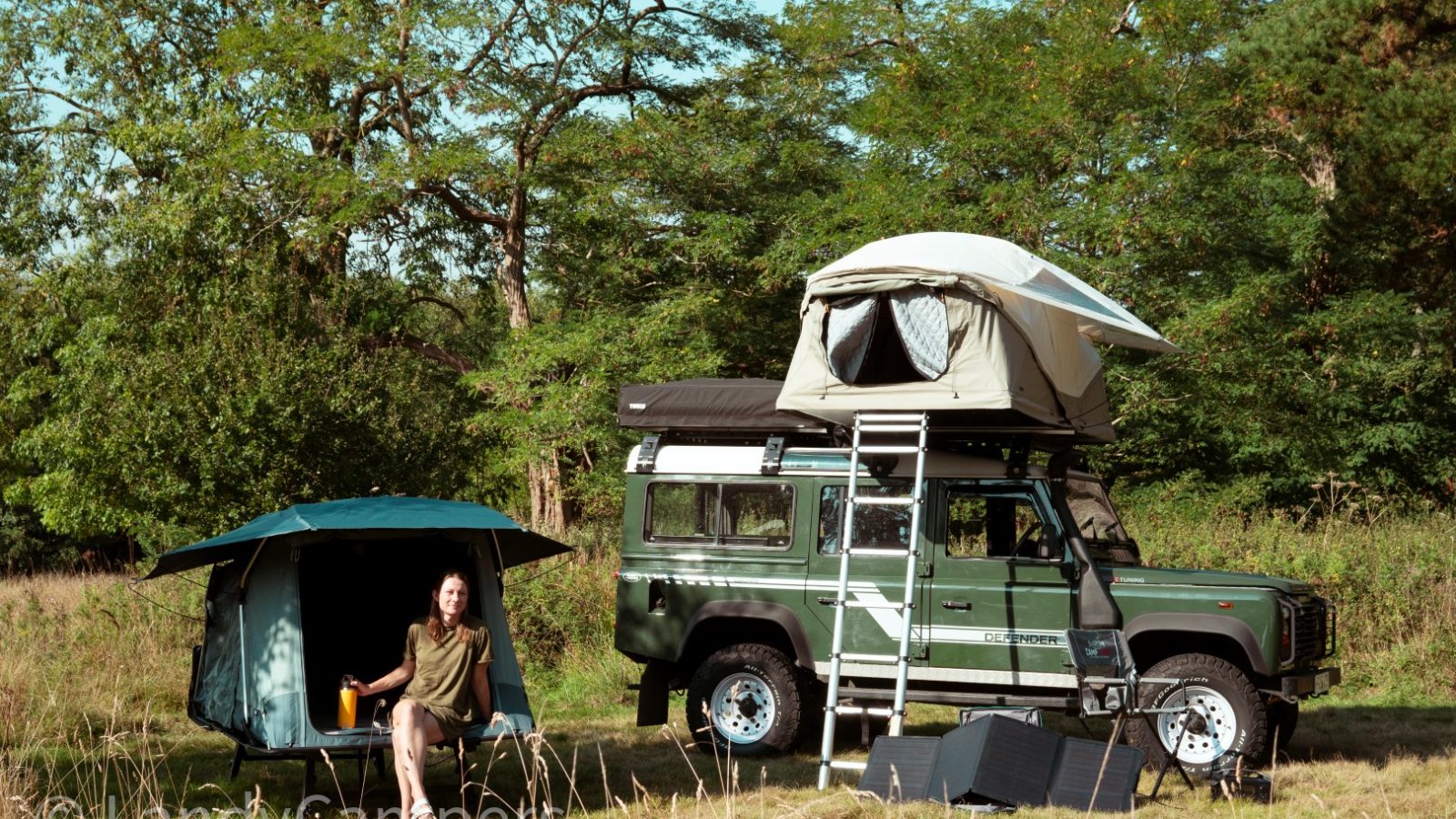 A woman relaxes by her Land Rover, a signature model from LandyCampers, complete with a roof tent. She's nestled in a grassy area surrounded by trees and camping gear, perfectly embracing the spirit of outdoor adventure.