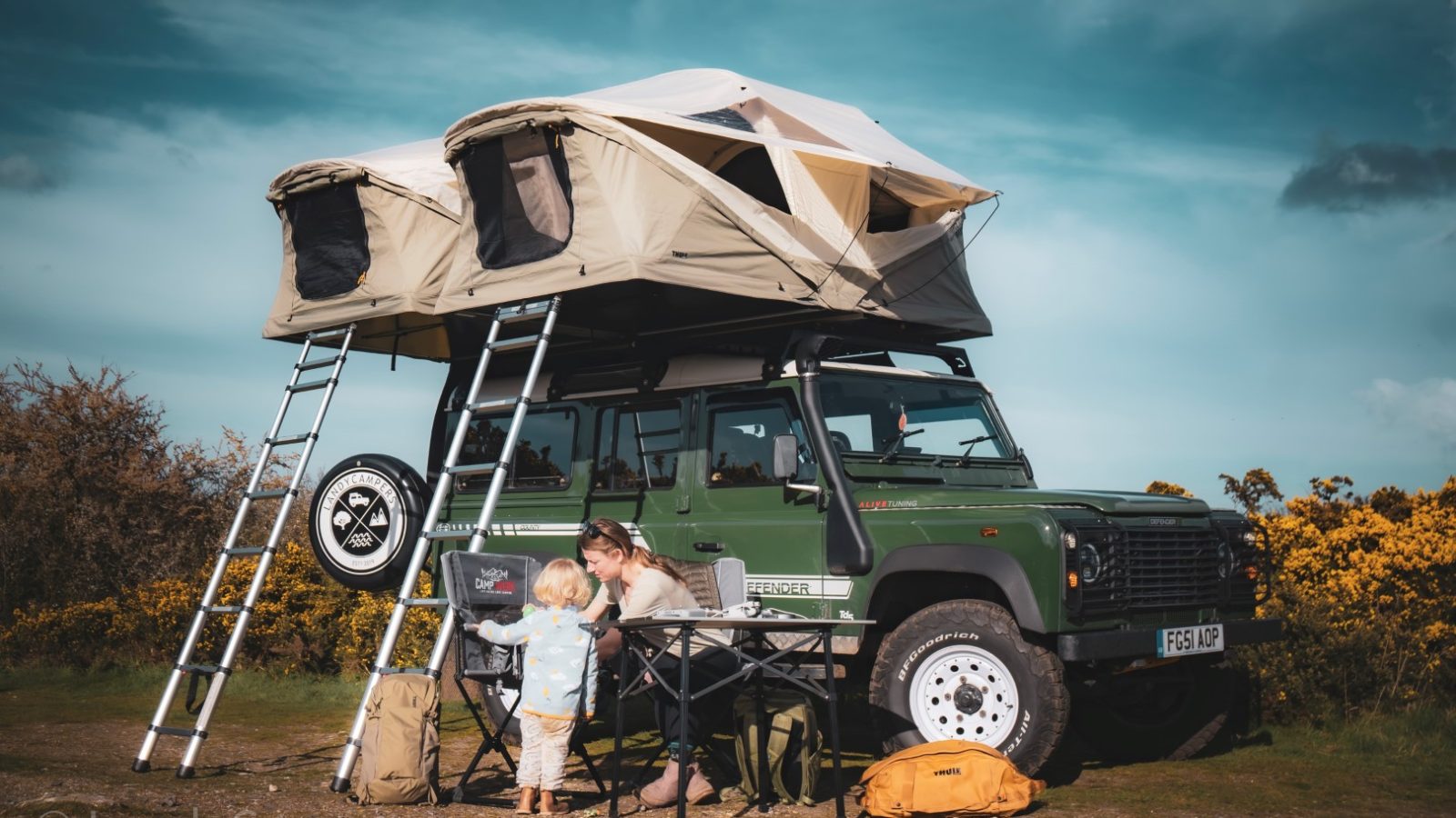 The family enjoys a sunny day at the campsite, their green Land Rover, kitted with LandyCampers essentials, stands proudly under a rooftop tent amid camping gear beneath the clear blue sky.