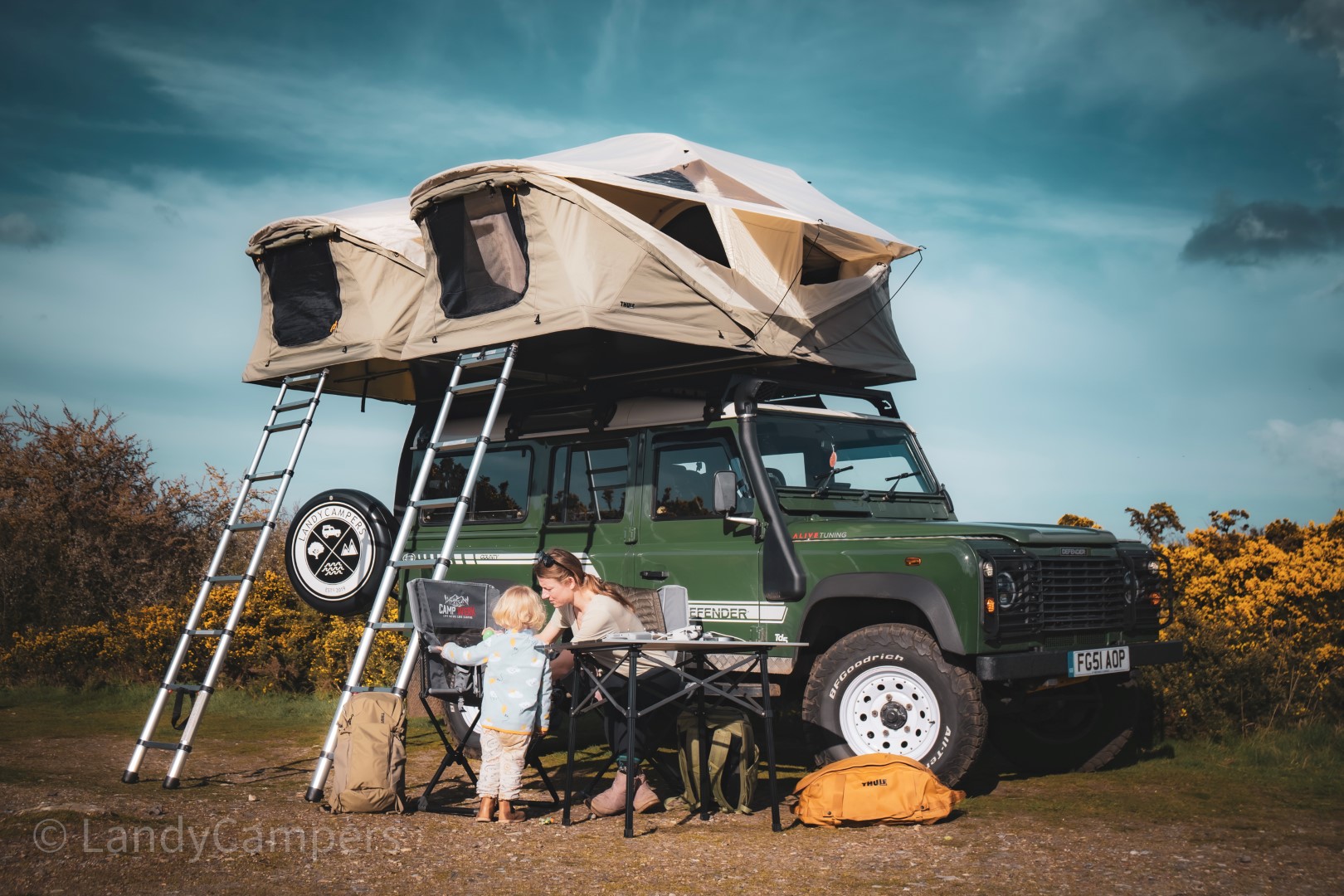 The family enjoys a sunny day at the campsite, their green Land Rover, kitted with LandyCampers essentials, stands proudly under a rooftop tent amid camping gear beneath the clear blue sky.