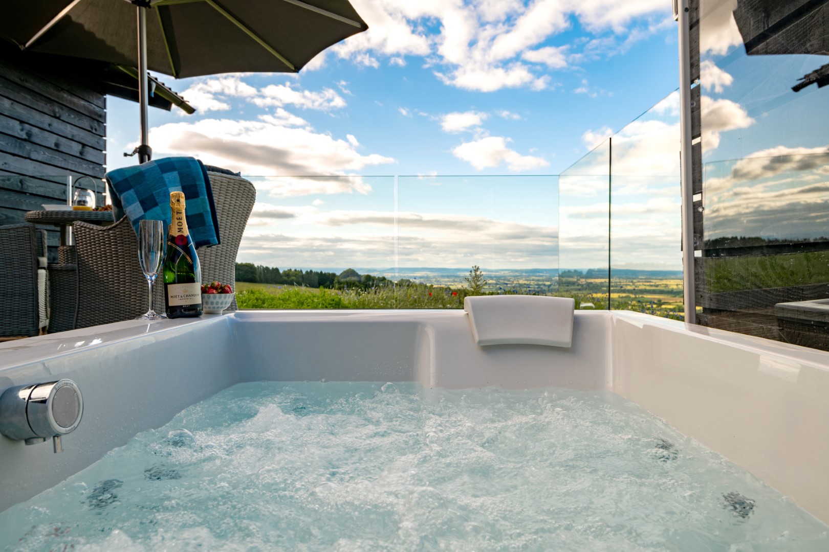 A hot tub with bubbling water overlooks a scenic landscape under a partly cloudy sky, perfect for an adults-only holiday, with a chair, umbrella, and champagne nearby.