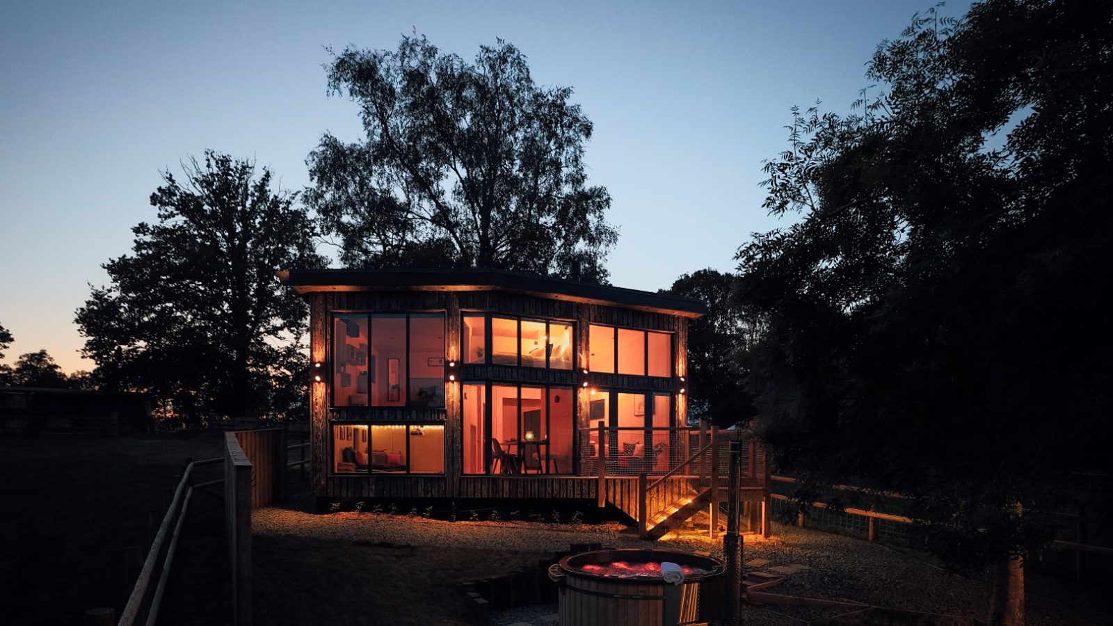 At dusk, the modern two-story glass house stands in harmony with nature, its warm lights glowing gently. Nestled among the trees, it offers a serene retreat at Orcop TreeTops, with a wooden hot tub enhancing the tranquil vibe in the yard.