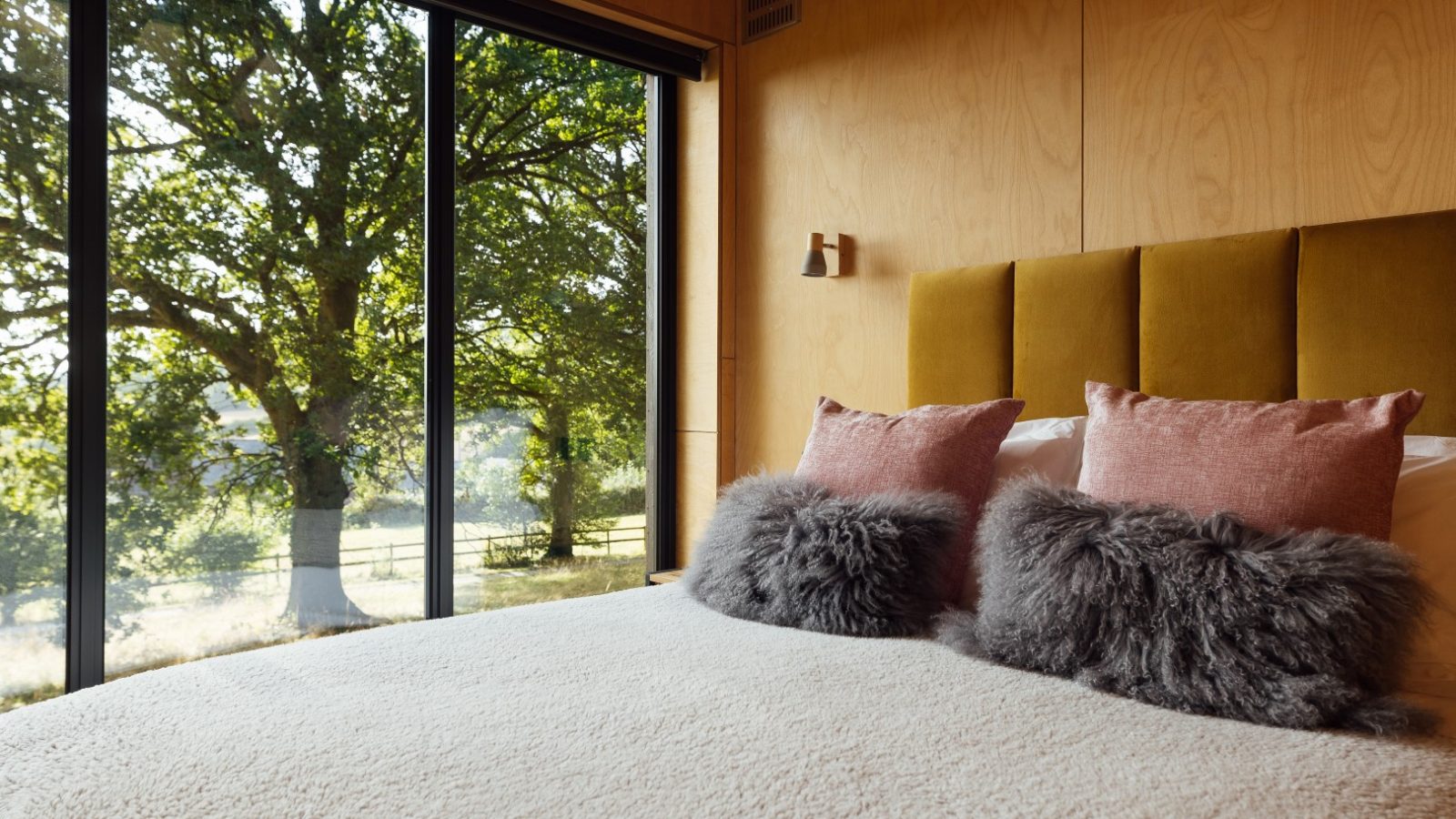 The cozy TreeTops bedroom features large windows, fluffy gray pillows, and a view of trees outside. With wood-paneled walls and a mustard headboard, it's the perfect retreat for nature lovers.