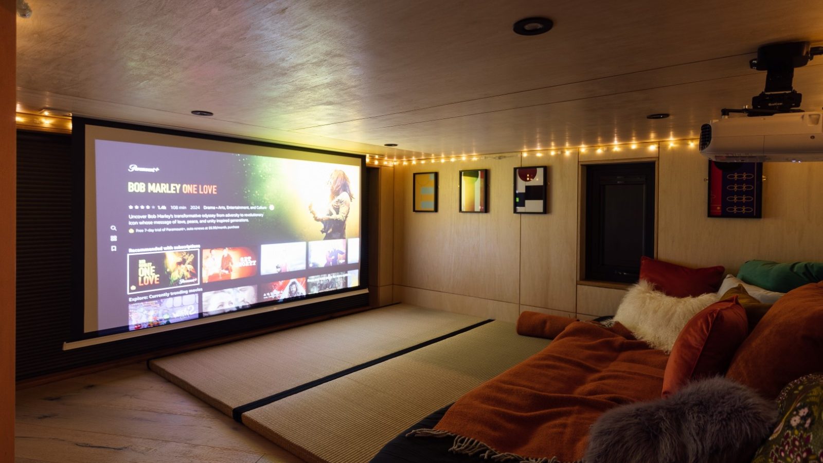 Cozy home theater with a large screen showing a Bob Marley documentary. Cushioned seating at Orcop TreeTops and string lights create a warm ambiance.