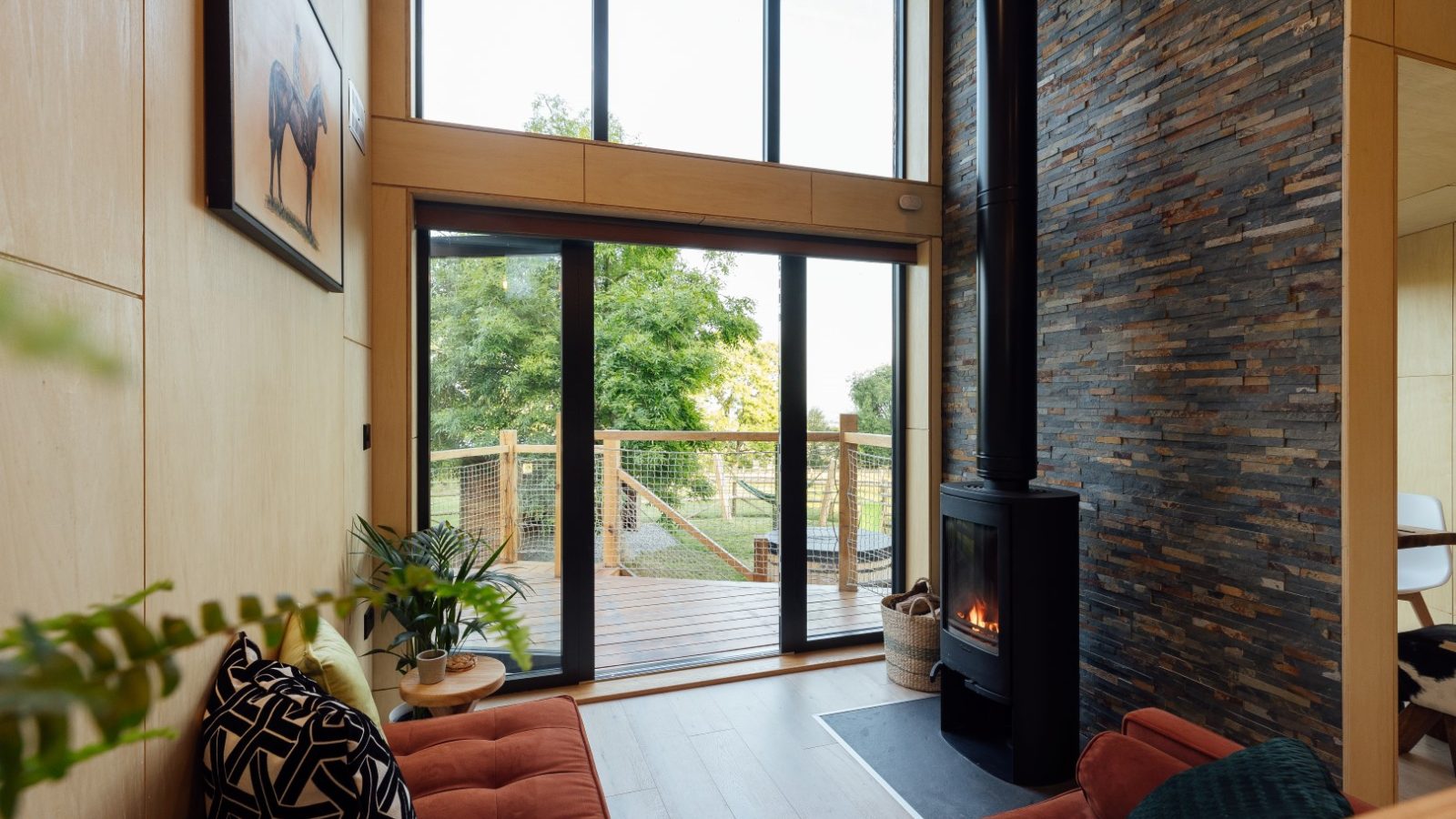 The modern living room features a stone fireplace and large windows that open up to a view of the wooden deck and surrounding TreeTops, creating a serene and inviting atmosphere.