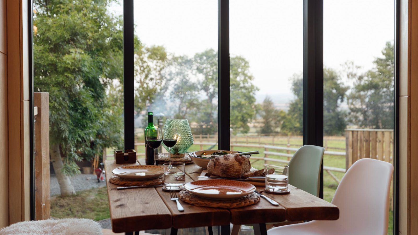 A wooden dining table set for dinner, adorned with wine glasses, plates, and bread, sits beside large windows that offer a breathtaking view of the lush landscape at Orcop TreeTops.