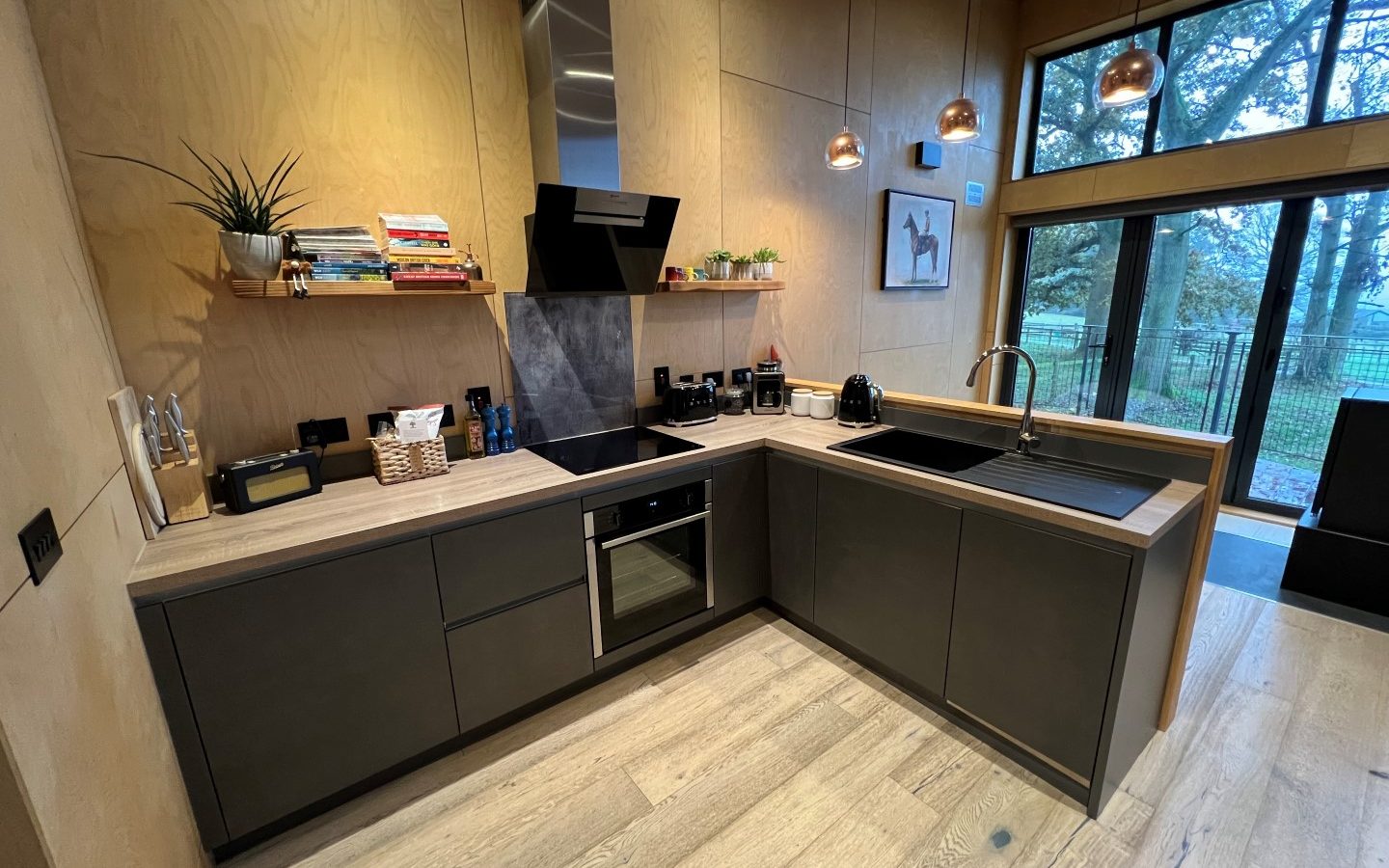 Modern kitchen with wood cabinetry, black appliances, and pendant lights. Large windows on the right overlook the lush greenery of Orcop TreeTops.