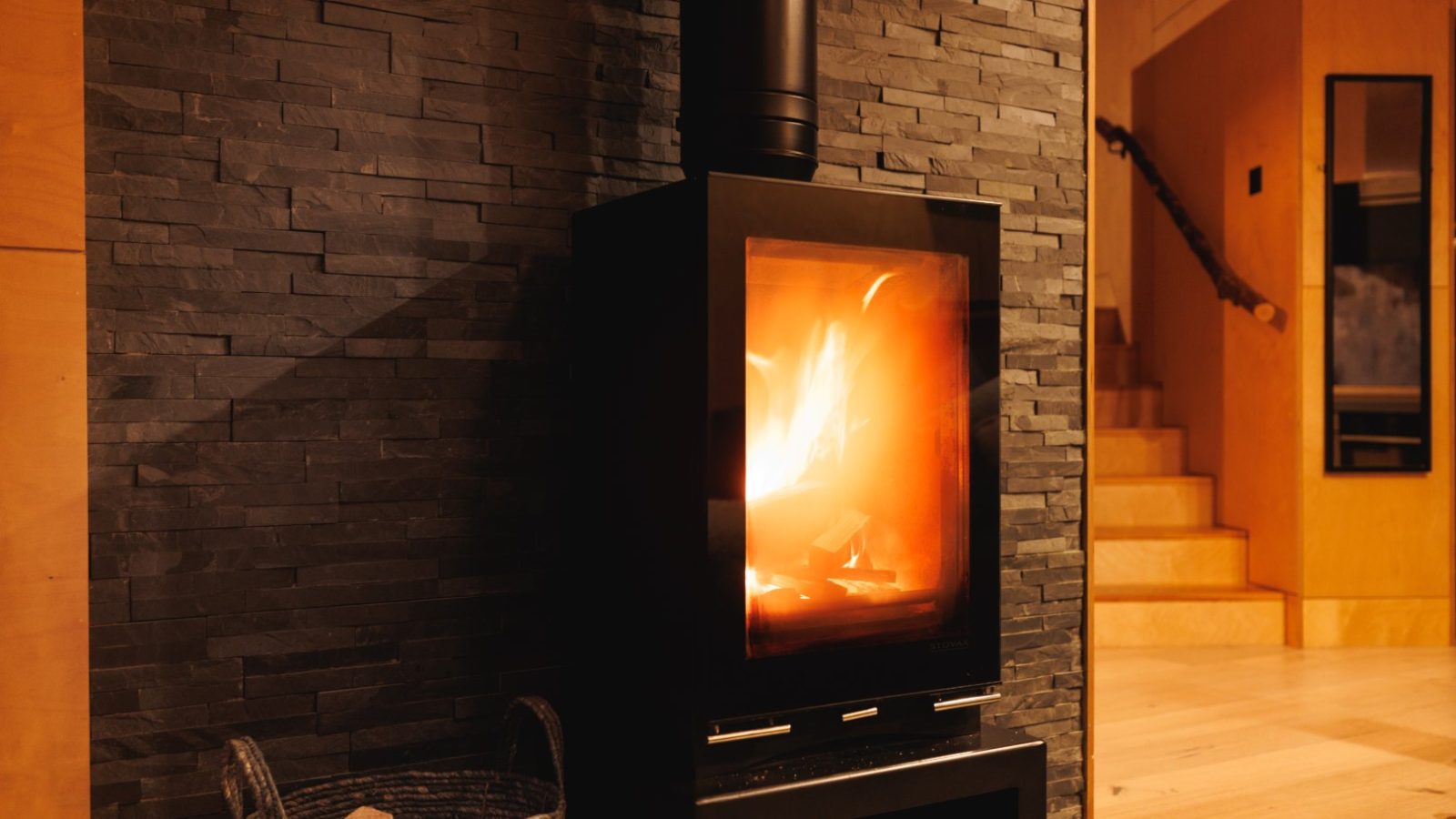 In the cozy room, an Orcop TreeTops black wood-burning stove crackles with flames, beautifully set against a dark stone wall. Nearby, a wicker basket completes the warm ambiance.