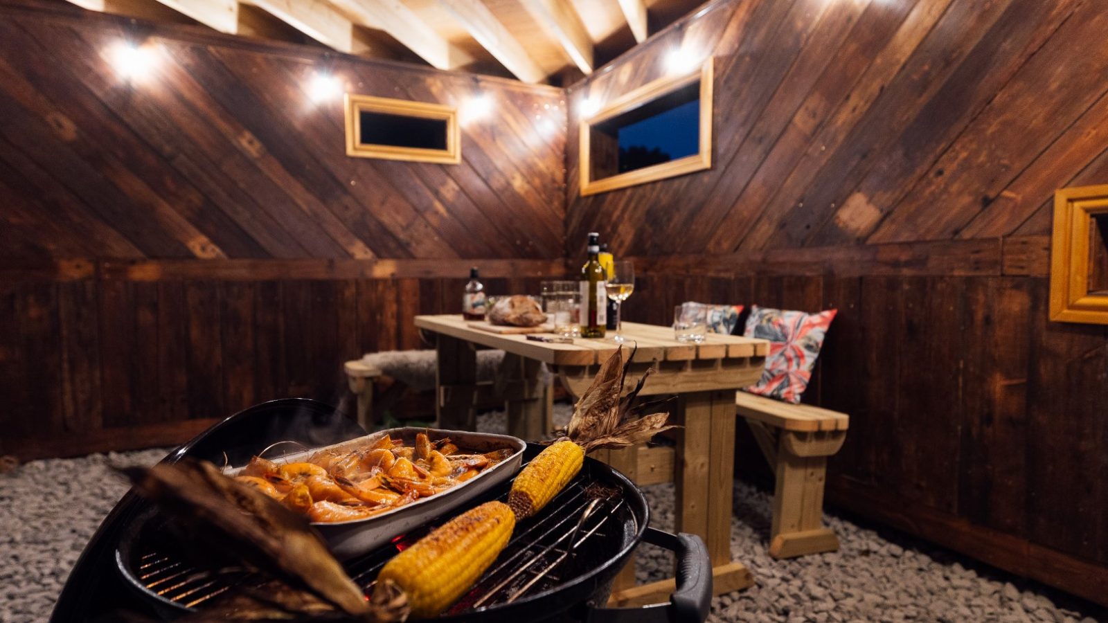 A barbecue grill with shrimp and corn nestles in an Orcop Tree patio area, featuring a table set with wine glasses and a cozy bench.