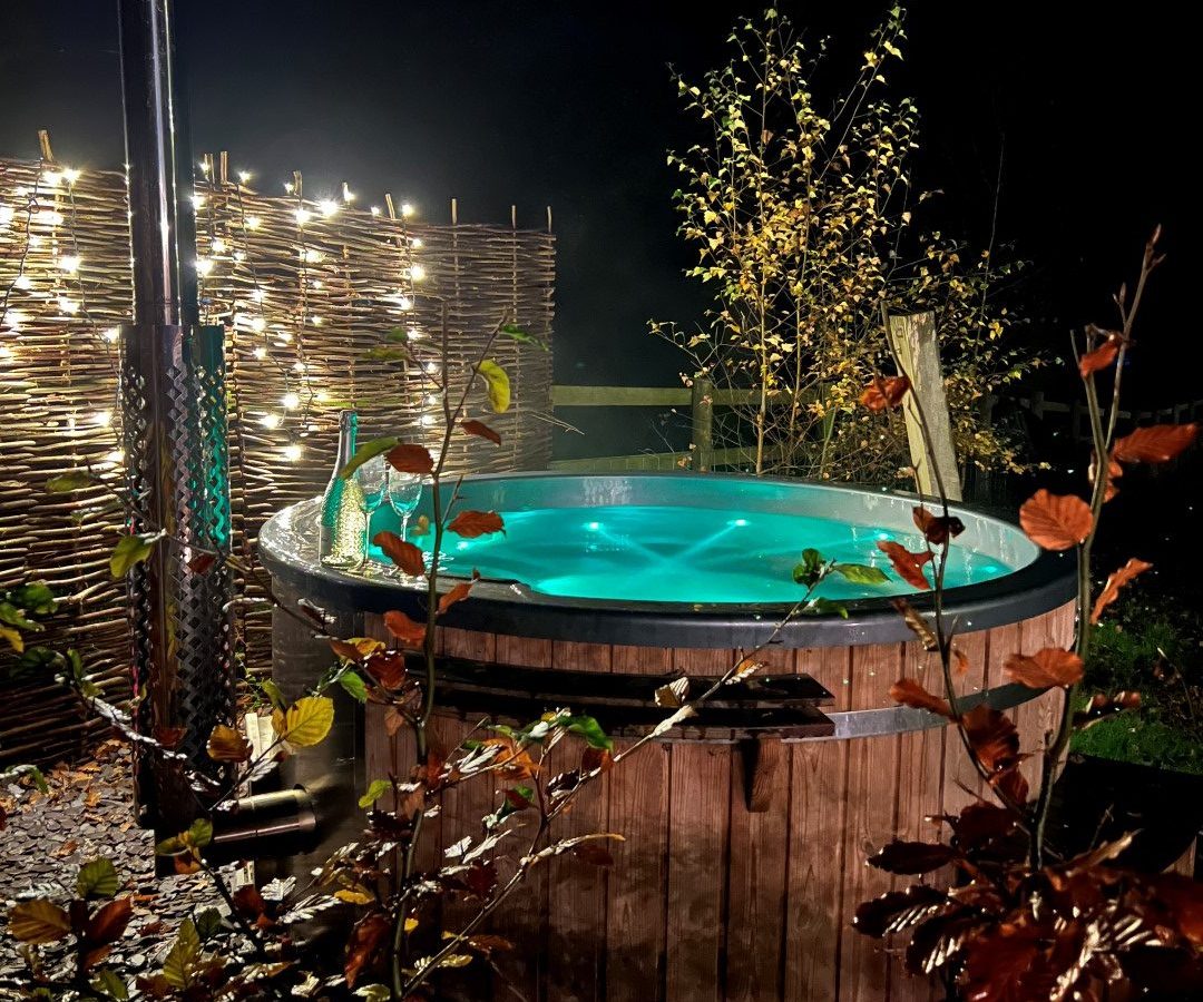 At the enchanting Orcop TreeTops, a wooden hot tub filled with water glows under the starlit sky, surrounded by fairy lights and lush foliage.