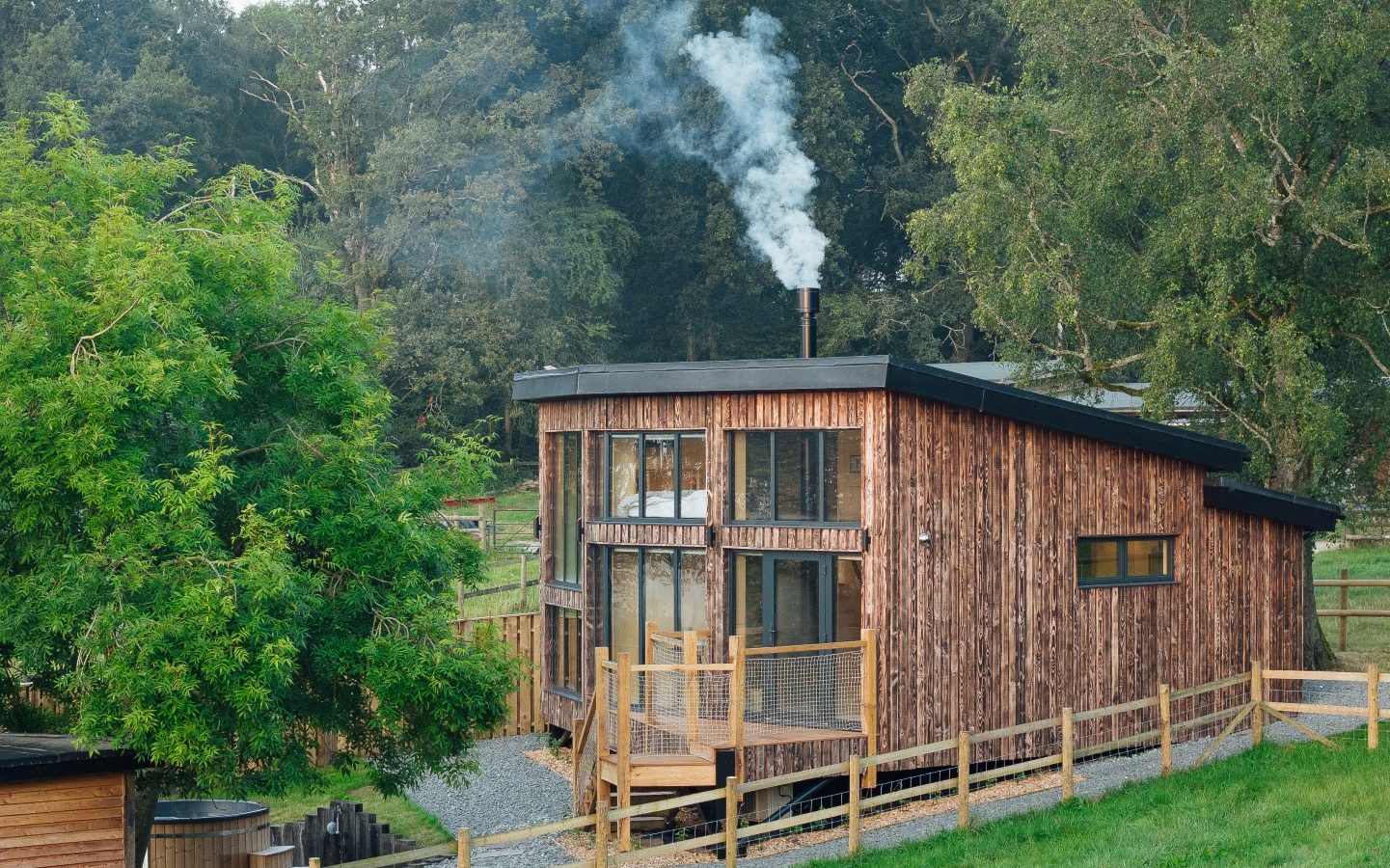 Nestled in a lush, green landscape, an Orcop cabin stands with smoke curling from the chimney. Surrounded by trees and a wooden fence, this serene retreat near the TreeTops offers a perfect escape into nature.