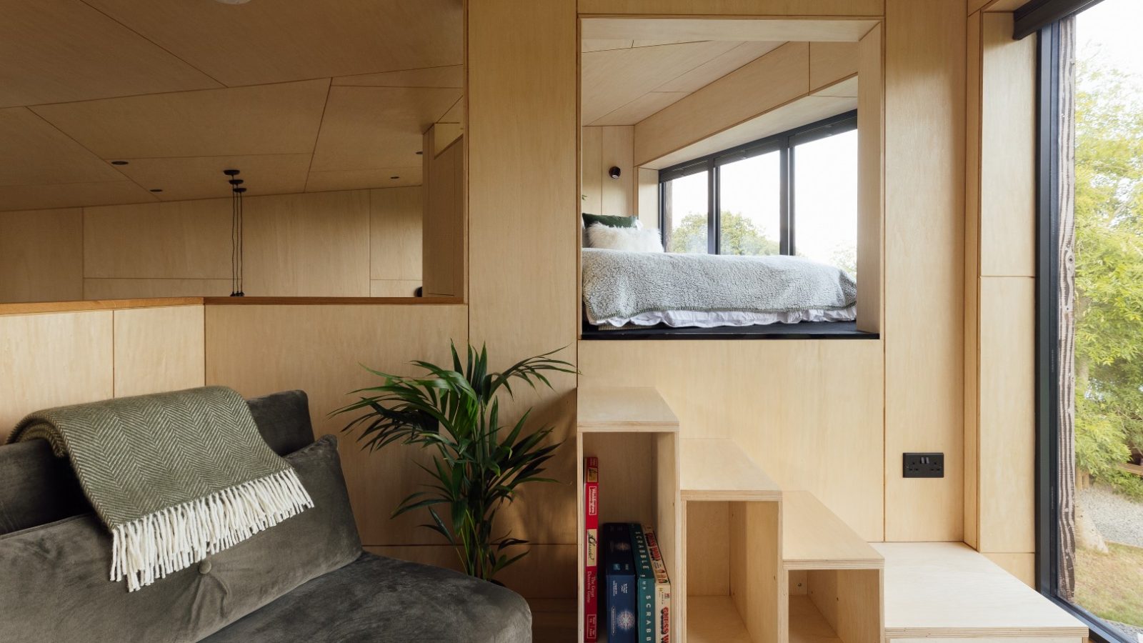 The interior of the modern Orcop TreeTops tiny house features a cozy loft bed, a window view, a gray couch, and a green plant. Wooden steps lead up with books neatly tucked beneath.