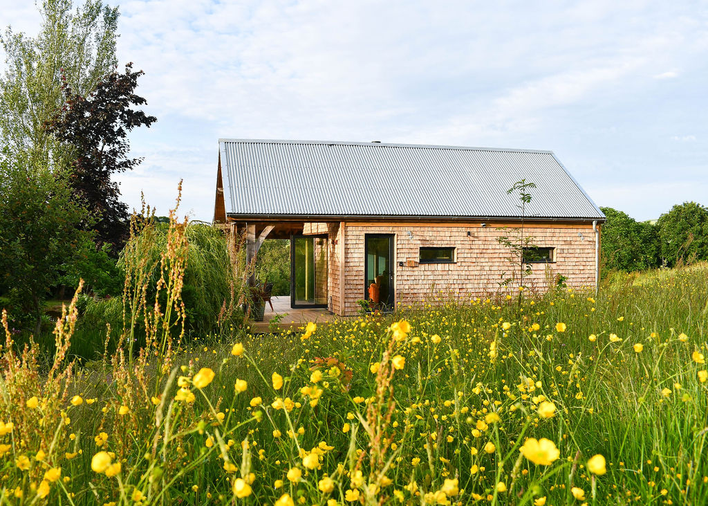 A quaint wooden house with a metal roof sits in a field of yellow wildflowers and grass under a partly cloudy sky, offering the perfect lodge break for an idyllic Easter getaway.