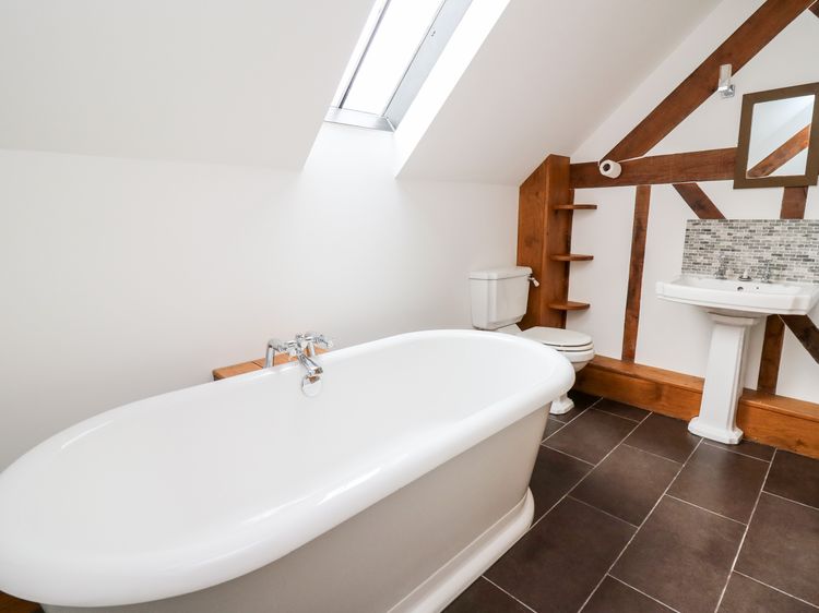 A modern bathroom with a freestanding bathtub, pedestal sink, and toilet is accentuated by skylights and wooden beam walls, offering a touch of charm reminiscent of Ploony Holidays in Wales.