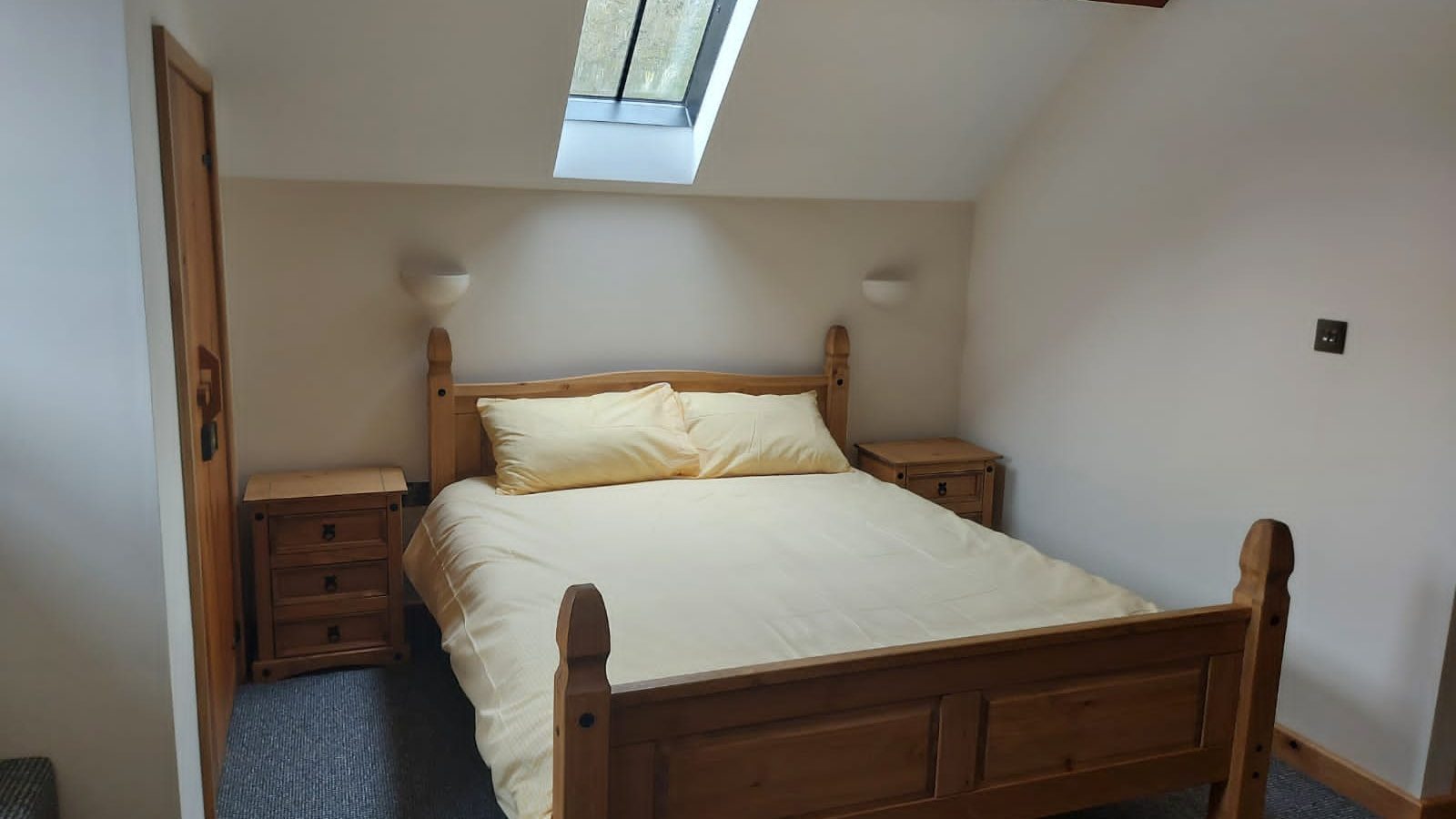 This cozy bedroom, ideal for a retreat in Wales, features a wooden bed with yellow bedding, two wooden nightstands, a carpeted floor, and a skylight window that bathes the room in natural light. Perfect for enjoying peaceful Ploony Holidays.